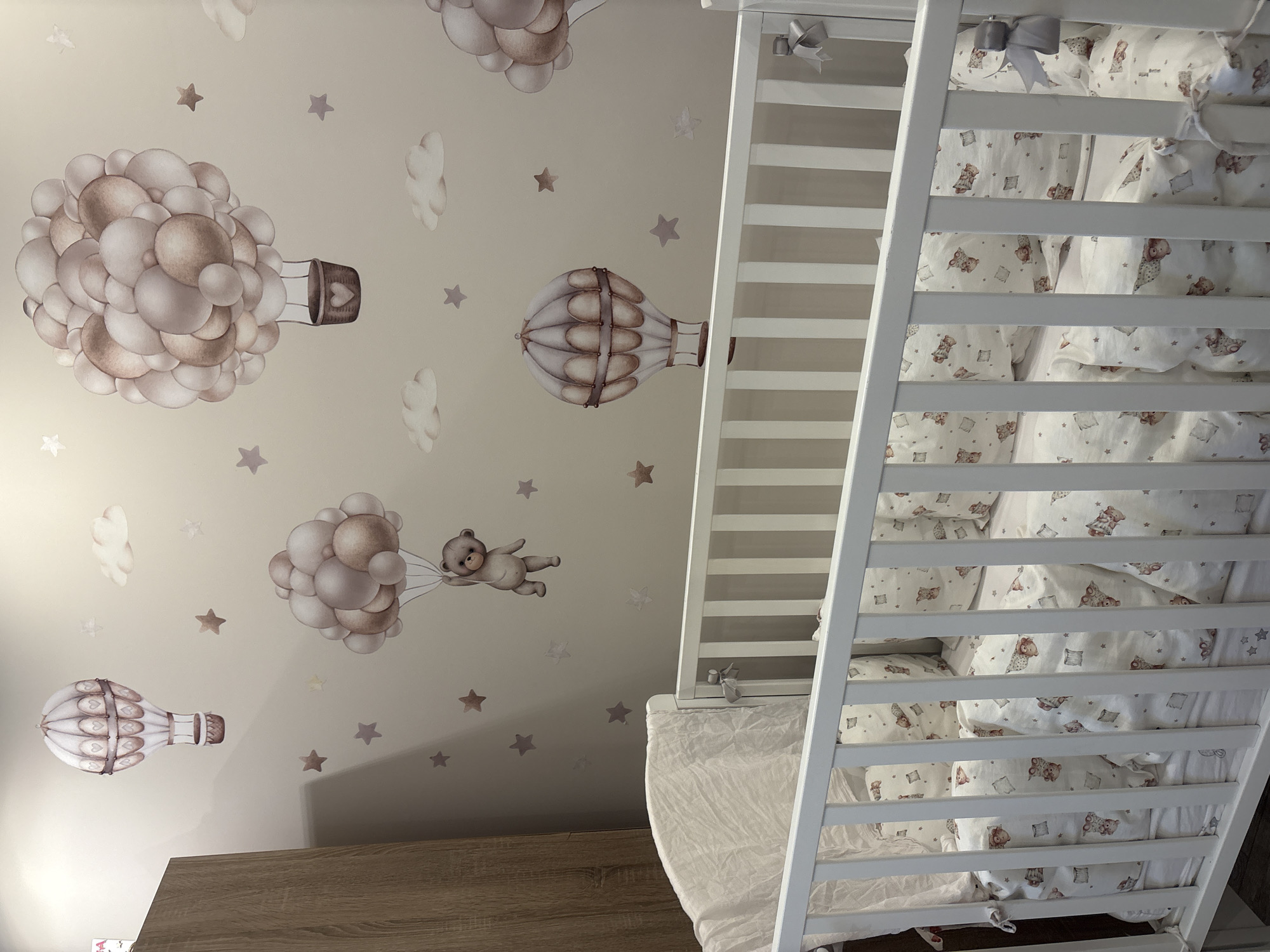 The image depicts a nursery room with a crib and a wall decorated with decals. The crib is white and appears to be made of wood, with white bedding featuring a pattern of teddy bears and stars. The wall behind the crib is painted a neutral color and is adorned with decals of hot air balloons, clouds, and stars in muted tones of brown and grey. One hot air balloon is particularly prominent, composed of many spherical shapes. Another decal shows a teddy bear holding onto balloons and floating in the sky. The scene is well-lit, suggesting it's daytime. The overall impression is one of a serene and whimsical nursery. No people are visible in the image. The location is identified as Strelcha, Bulgaria. There is no visible text in the image.