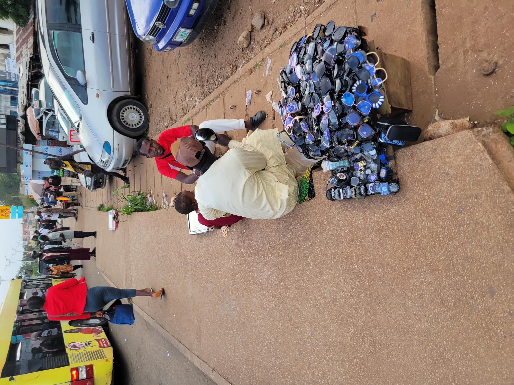 A street vendor in Jos, Nigeria, sits on a sidewalk next to a large pile of sunglasses, likely for sale. He holds one pair up to his eye. Beside him, another person, possibly a customer, watches him. The sidewalk is lined with a variety of watches and other small items, also for sale. A bus and several cars are parked along the street, indicating a busy urban environment. The scene suggests a typical market day in Jos, with vendors and shoppers interacting in a bustling commercial area.