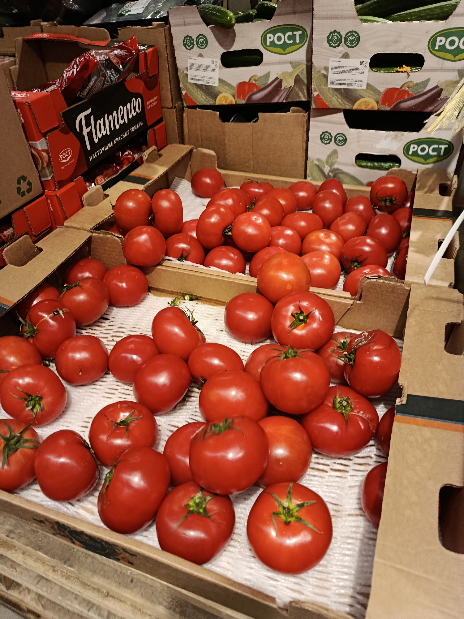 The image shows a display of ripe, red tomatoes in cardboard crates at a market stall, possibly in Orekhovo-Zuyevo, Russia. Several crates are stacked, with the tomatoes arranged in rows on a white, textured surface. Some of the crates have branding, including the word "Flamenco" and the logo "РОСТ". In the background, there are also crates of cucumbers and what appear to be eggplants and peppers. The overall impression is of a fresh produce section in a retail environment.