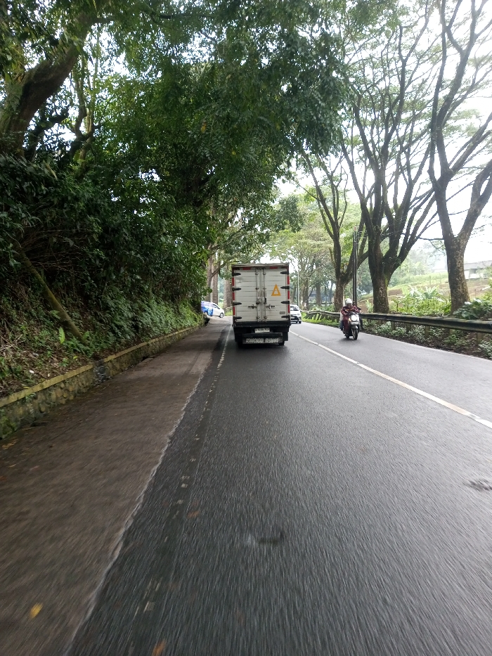 The image depicts an outdoor scene on a two-lane asphalt road in the city of YonPhoogla, Bhutan, under overcast conditions. The road surface is wet, suggesting recent precipitation.
In the foreground, the perspective is from a moving vehicle, facing forward. A solid white line delineates the left edge of the road, separating it from a paved shoulder or path. A stone retaining wall is visible along the base of a heavily vegetated embankment on the far left. Large trees with dense green foliage line this embankment, overhanging the road.
In the midground, a white box truck with two yellow triangular hazard symbols on its rear doors is positioned in the left lane, moving away from the camera. Its license plate, partially visible, appears to be a white plate with black characters. To the right of the truck, a person wearing a white helmet and a reddish-brown jacket is operating a white scooter in the right lane, also moving away. A solid yellow line separates the two lanes.
Further down the road, in the background, a white car, possibly a sedan, is parked or stopped on the paved shoulder to the left of the truck. Another white car, possibly an SUV, is visible in the right lane beyond the scooter. A metal guardrail runs along the right side of the road, and beyond it, additional trees with sparser foliage and distant hills are visible. The sky is uniformly light and overcast.