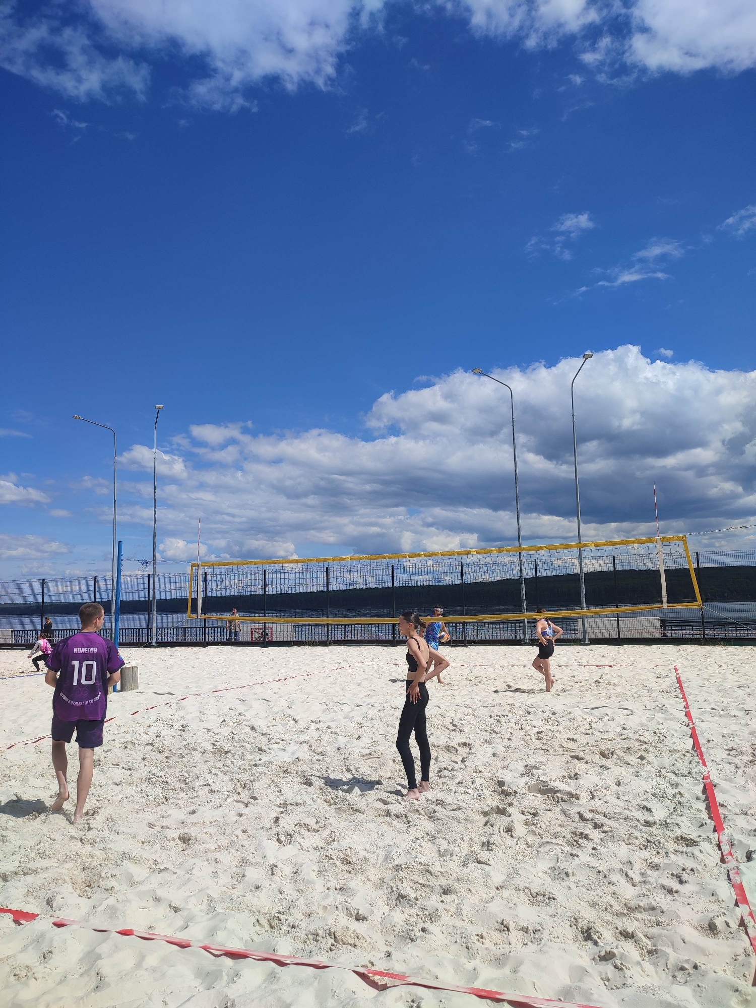 The image captures a beach volleyball court on a sunny day at an unknown location in Unknown, Unknown. The foreground features a white sand court, partially delineated by red and white striped boundary tape. Five individuals are present on or near the court. In the left foreground, a male subject is viewed from behind, wearing a purple athletic shirt with "КОЛЕГОВ" and the number "10" printed on the back, along with purple shorts, walking barefoot. To his right, a female subject stands barefoot in a black sports bra and black leggings, facing right with hands on hips. Further right and slightly behind the net, another female subject in a patterned sports bra and black shorts stands barefoot. A fourth person, partially visible behind the first female subject, holds a yellow volleyball. A fifth individual is seen bending over in the far left background. The court is equipped with a yellow and black volleyball net. Behind the net, two tall streetlights with rectangular LED luminaires are prominent. A dark metal fence separates the court area from a large body of water, beyond which a distant, dark forested landmass is visible. The sky is bright blue with scattered white cumulus clouds.