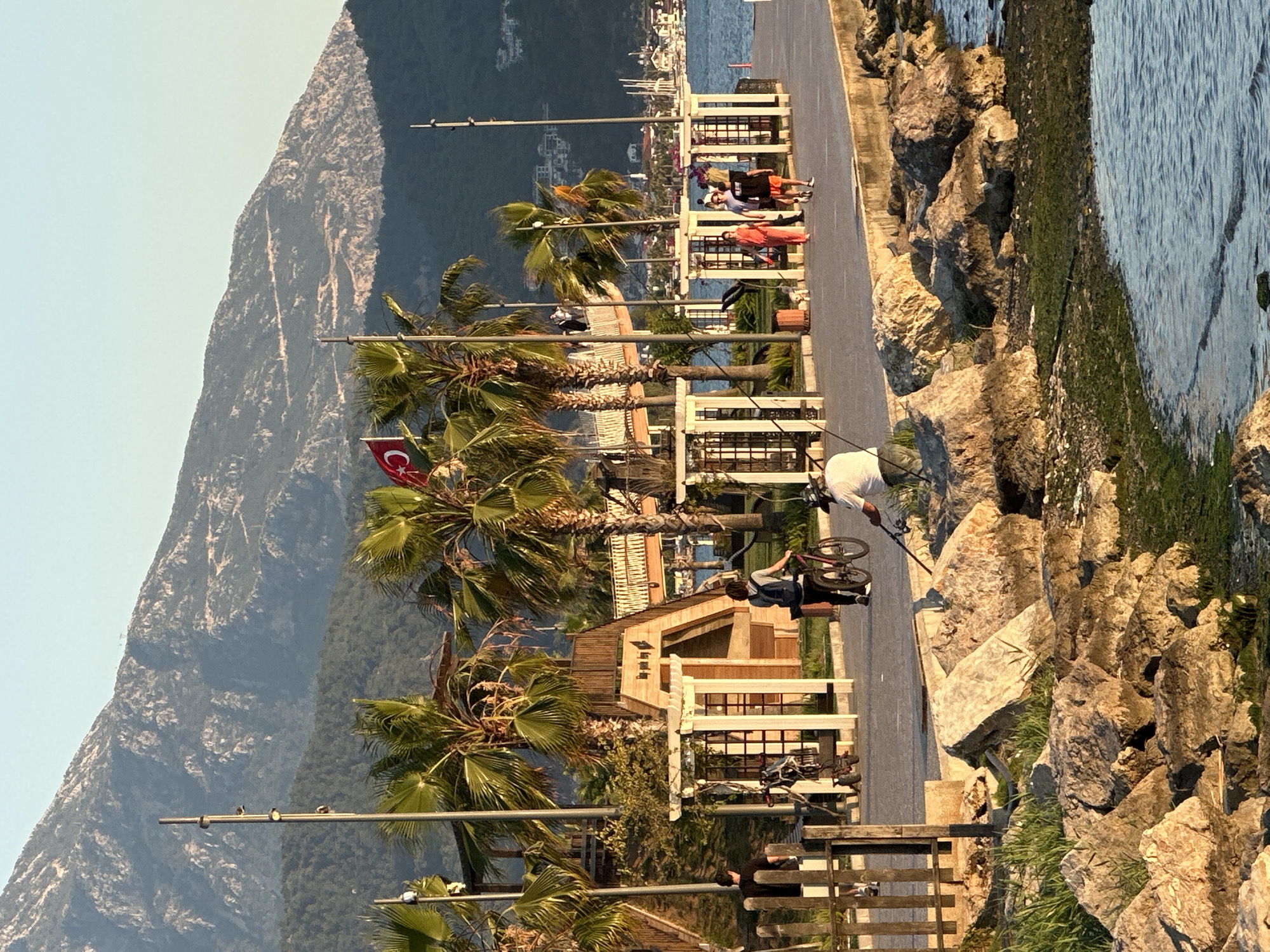 A picturesque coastal scene in Fethiye, Turkey, captures a lively promenade running parallel to the sea, bathed in the warm, golden light of late afternoon or early evening. The paved promenade, positioned on the left, is adorned with numerous tall palm trees and streetlights, some of which feature birds perched atop them. White fences line the path, behind which several small wooden structures, possibly cafes or kiosks, are visible.
Several people are enjoying the waterfront. One individual is actively fishing from the rugged, moss-covered rocky edge of the water on the right, while another is walking alongside a bicycle on the promenade. Other figures are seated at outdoor tables, contributing to a relaxed atmosphere. A distinctive Turkish flag, featuring a white star and crescent on a red background, is prominently displayed on a streetlight, signaling the location.
To the right, the tranquil sea meets the rocky shoreline. The water's surface reflects the soft light, and distant boats are visible on the horizon. In the background, majestic, lush green mountains dominate the horizon under a clear, light blue sky. Hints of a distant town or settlement can be seen across the water. The overall impression is one of a calm, leisurely coastal setting under clear, sunny skies.