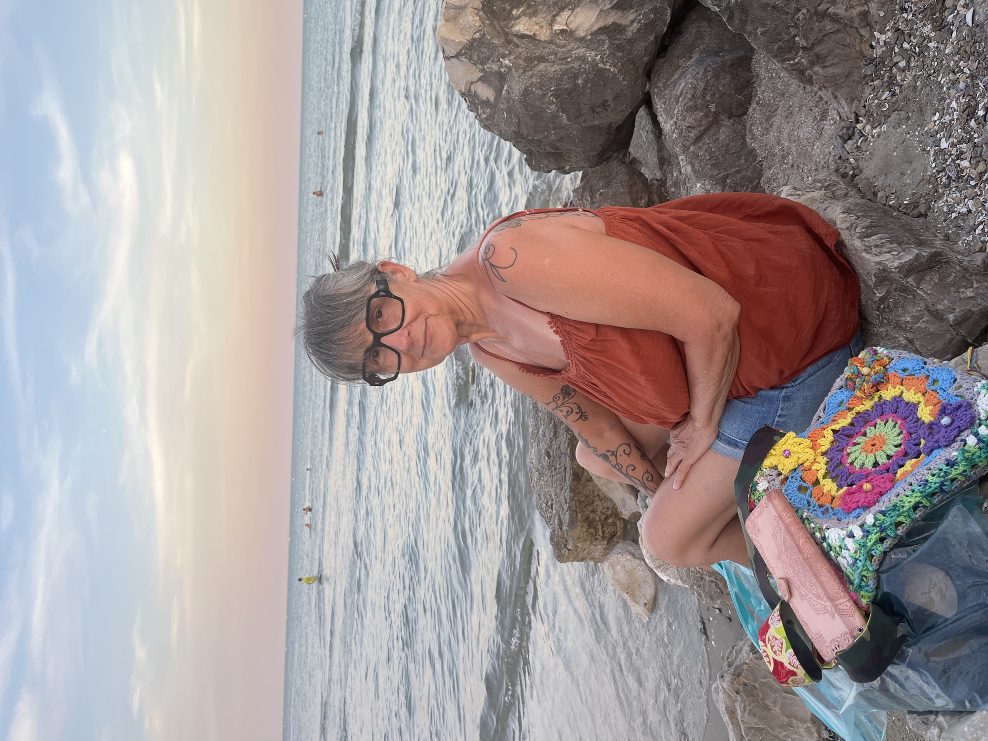 A middle-aged woman with gray hair and glasses sits on rocks by the sea. She is wearing an orange top and denim shorts. In front of her, there are several bags, including a colorful crocheted bag. The sea is calm with a few people swimming in the distance. The sky is painted with pastel hues of pink and blue, indicating either sunrise or sunset. The location is a beach in Palavas-les-Flots, France, with large rocks on the shore.
