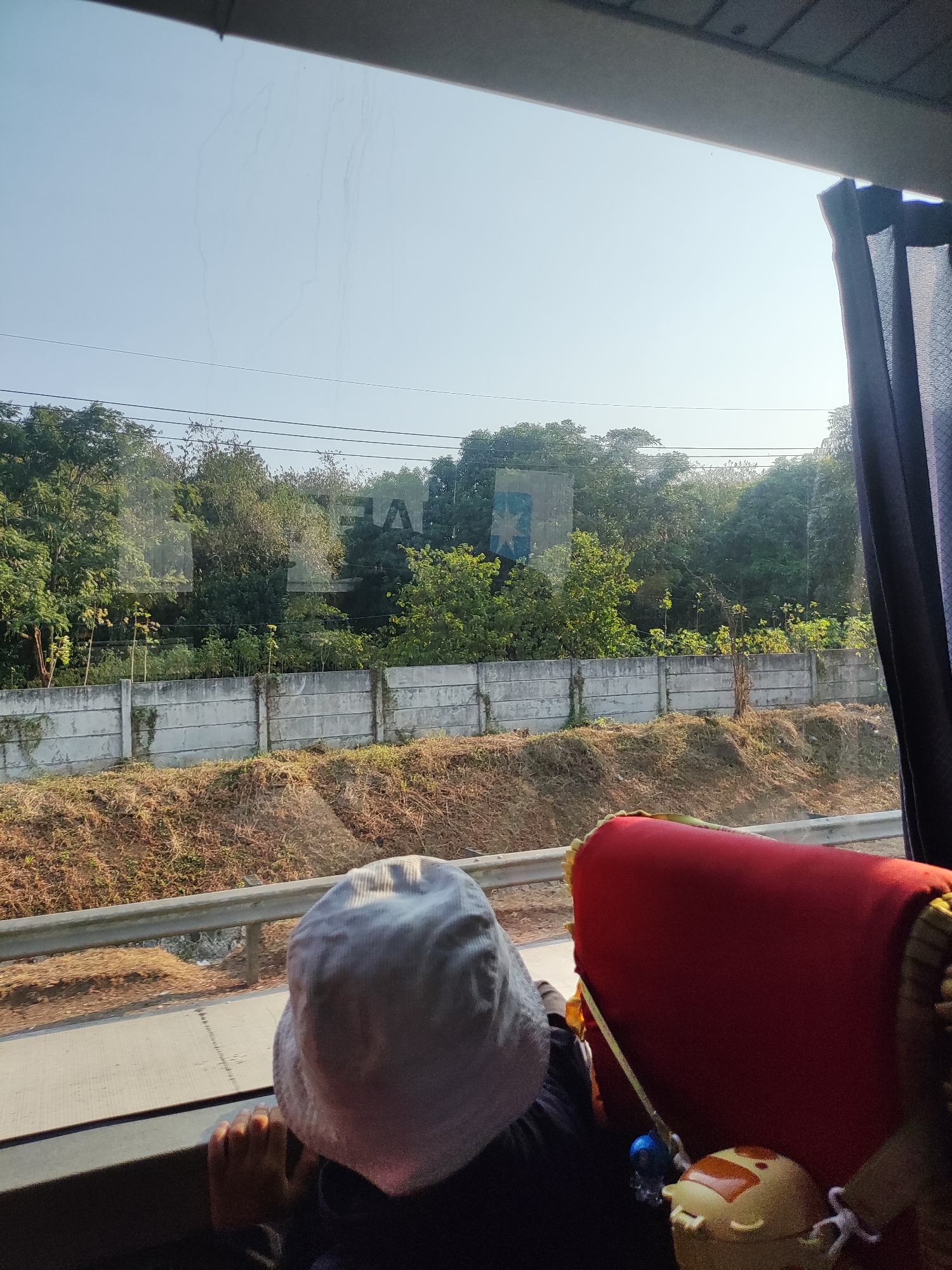 The image captures a view from inside a vehicle, likely a bus, looking out a window. In the foreground, a child wearing a light-colored bucket hat is looking out the window, resting their hands on the sill. To the right, a red seat with yellow trim is visible, and a small, yellow stuffed toy with an orange face is resting on it.
Through the window, a concrete wall stretches horizontally across the middle ground, with overgrown vegetation on either side. Behind the wall, lush green trees and foliage form a dense backdrop. The sky above is clear and bright, with faint, wispy streaks that could be contrails. A large, rectangular sign with a blue and white logo, possibly a shipping company like Maersk, is visible through the trees. The environment suggests a rural or semi-rural setting, possibly on the outskirts of Karawang, Indonesia, given the presence of industrial or logistical signage and the tropical vegetation. The overall scene conveys a sense of travel and observation.
