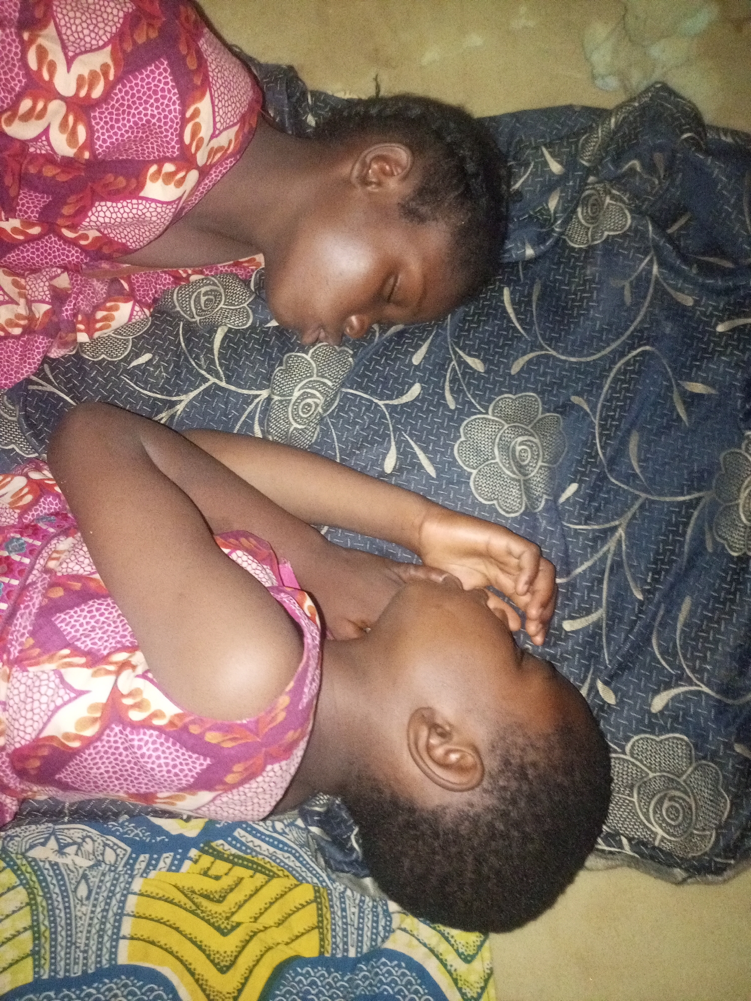 In Gombe, Nigeria, two young children are lying on a mat. One child is on their back, with their head turned to the left, and the other child is lying on their stomach, also with their head turned to the left. Both children are wearing patterned fabric, one with pink, purple, and orange designs, and the other with a dark blue fabric featuring white floral patterns. The background is a textured surface, possibly a floor or wall, with a yellowish-brown hue. The lighting is dim, suggesting an indoor setting. The scene appears to be a domestic environment.