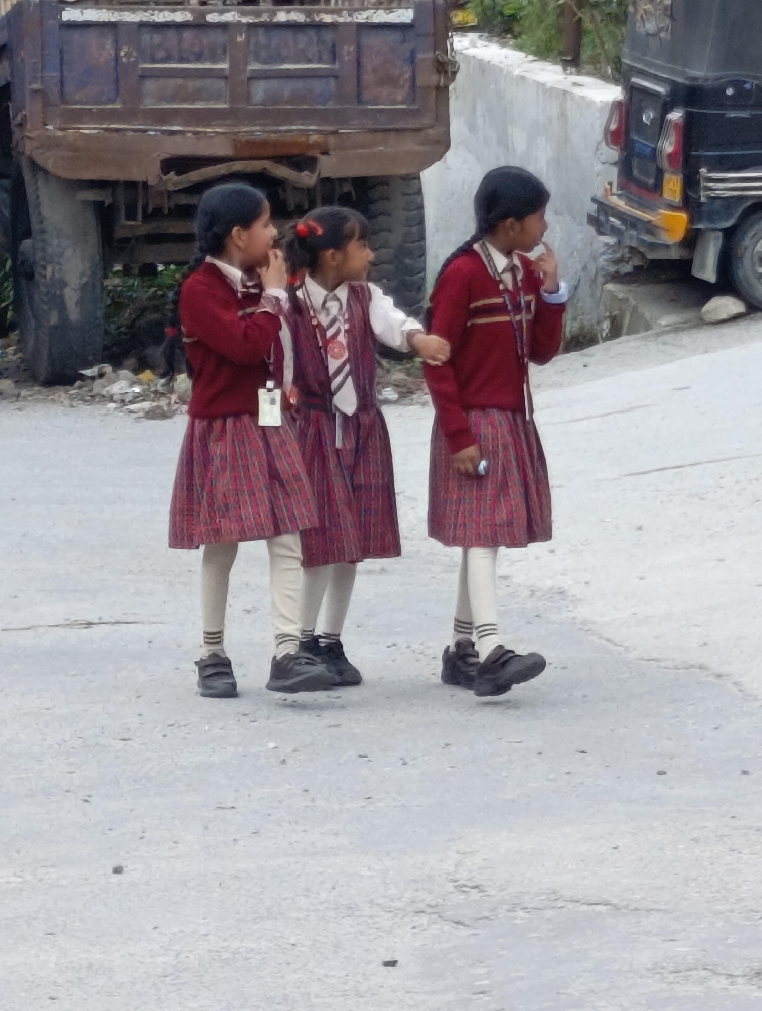 In Chhyal, India, three young schoolgirls, identified by their matching uniforms, stand on a paved road. The girls are wearing pleated plaid skirts, white stockings, and dark shoes. The girl on the left has a maroon sweater over a white shirt, and the girl in the middle wears a maroon vest over a white shirt with a striped tie. The girl on the right is also wearing a maroon sweater over a white shirt with a striped tie. In the background, a rusty truck is parked to the left, and a dark-colored auto-rickshaw is visible to the right. The environment appears to be a roadside setting with a concrete barrier separating the road from some vegetation behind the auto-rickshaw.