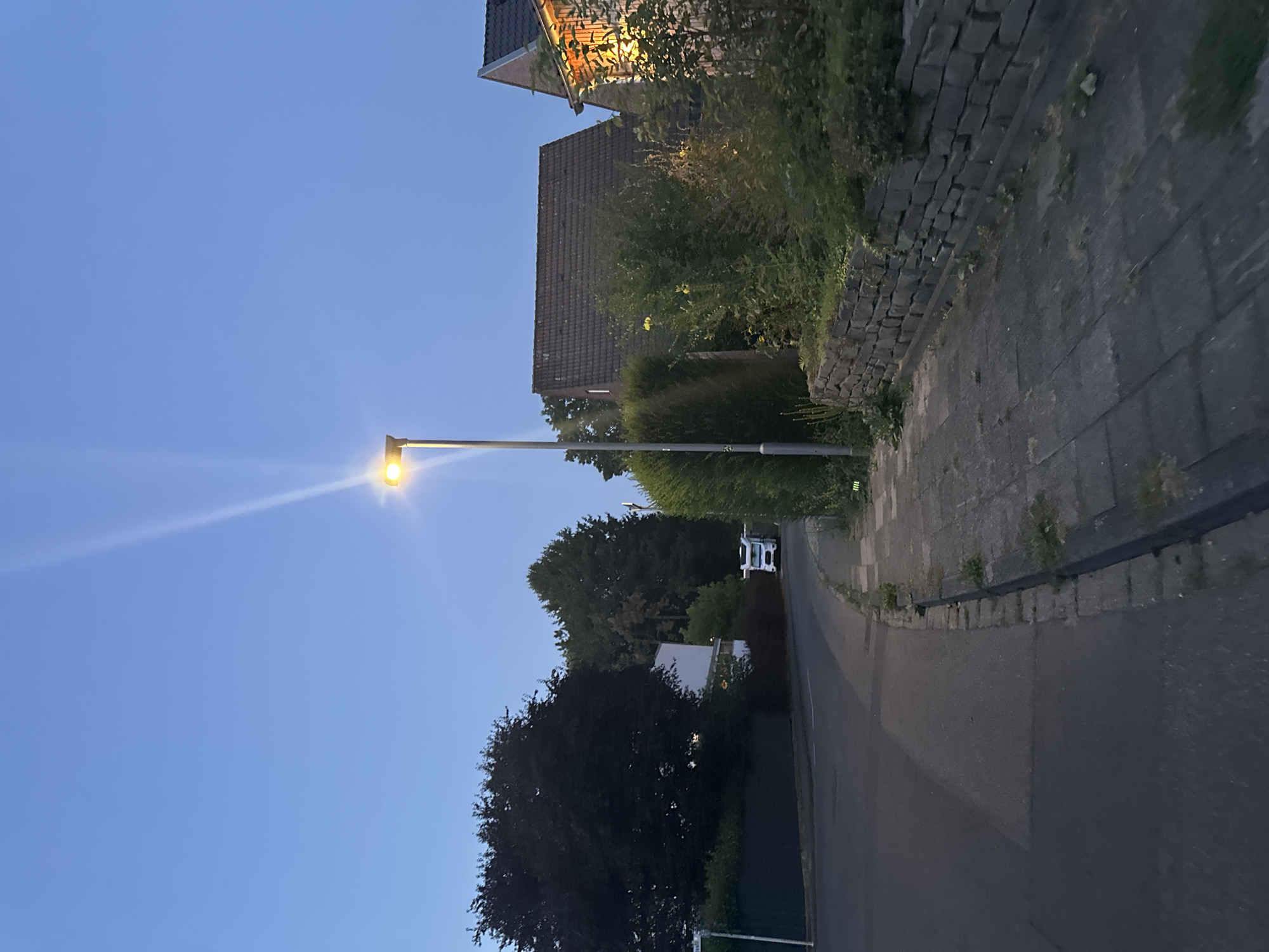 The image depicts a street scene in Krefeld, Germany, at dusk or early evening. A street lamp illuminates the area, casting a bright light upwards and to the right. On the right side of the street, a stone wall separates the sidewalk from a residential property with a house visible, its windows lit from within, suggesting habitation. The sidewalk is paved with concrete slabs and shows some patches of grass growing through the cracks. On the left side of the street, a road curves slightly to the left, and beyond it, a large, dark-leaved tree dominates the foreground. Further down the road, a white camper van is parked. The sky is a clear, deep blue, indicative of twilight. The scene appears peaceful and residential.
