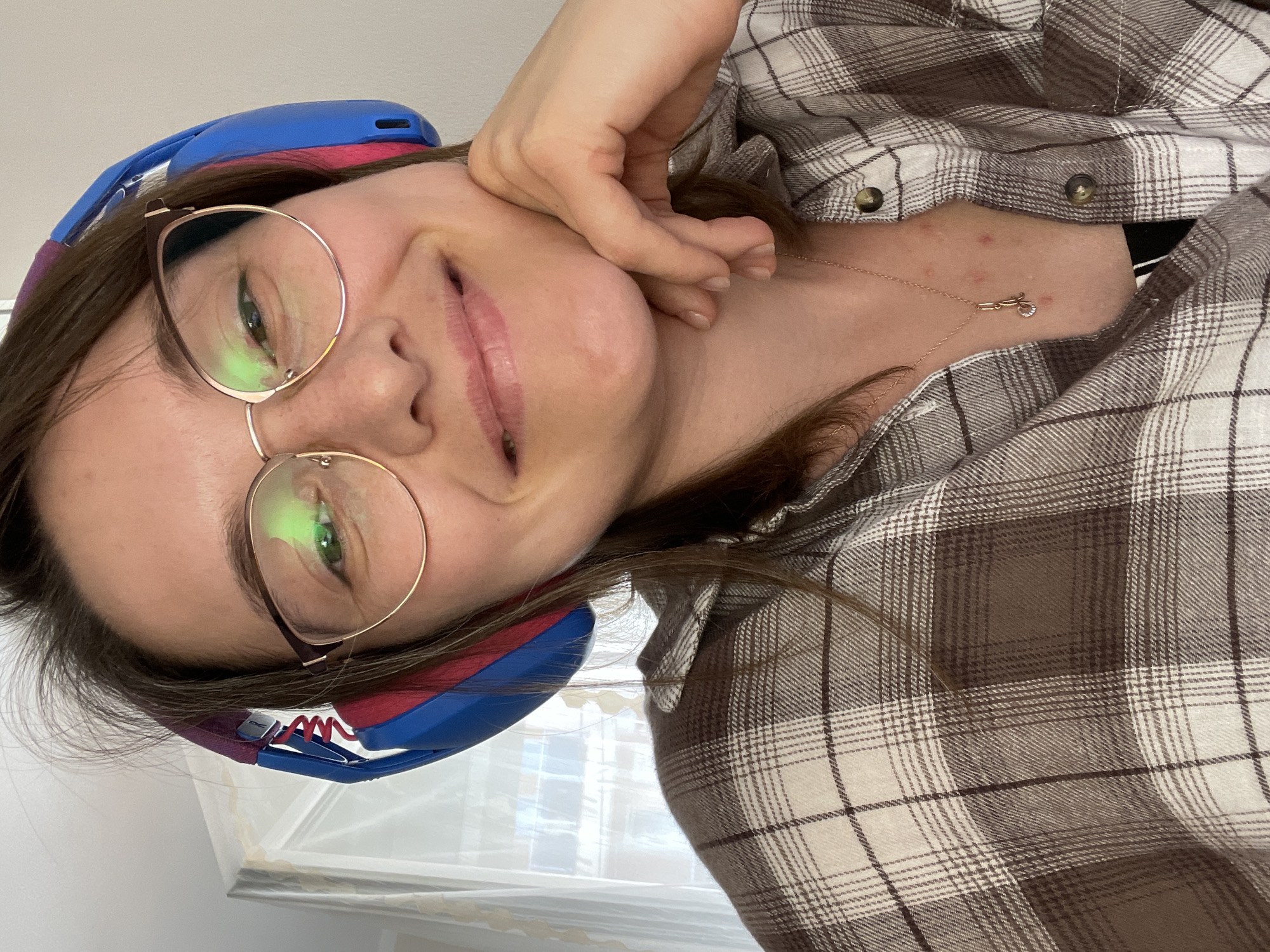 A woman wearing glasses and headphones is posing for a selfie in an indoor setting. She is wearing a plaid shirt and a necklace with a small charm. Her hand is near her face. The location is likely not in Budva, Montenegro, as there is no evidence of it in the image. The background includes a window with sheer curtains.