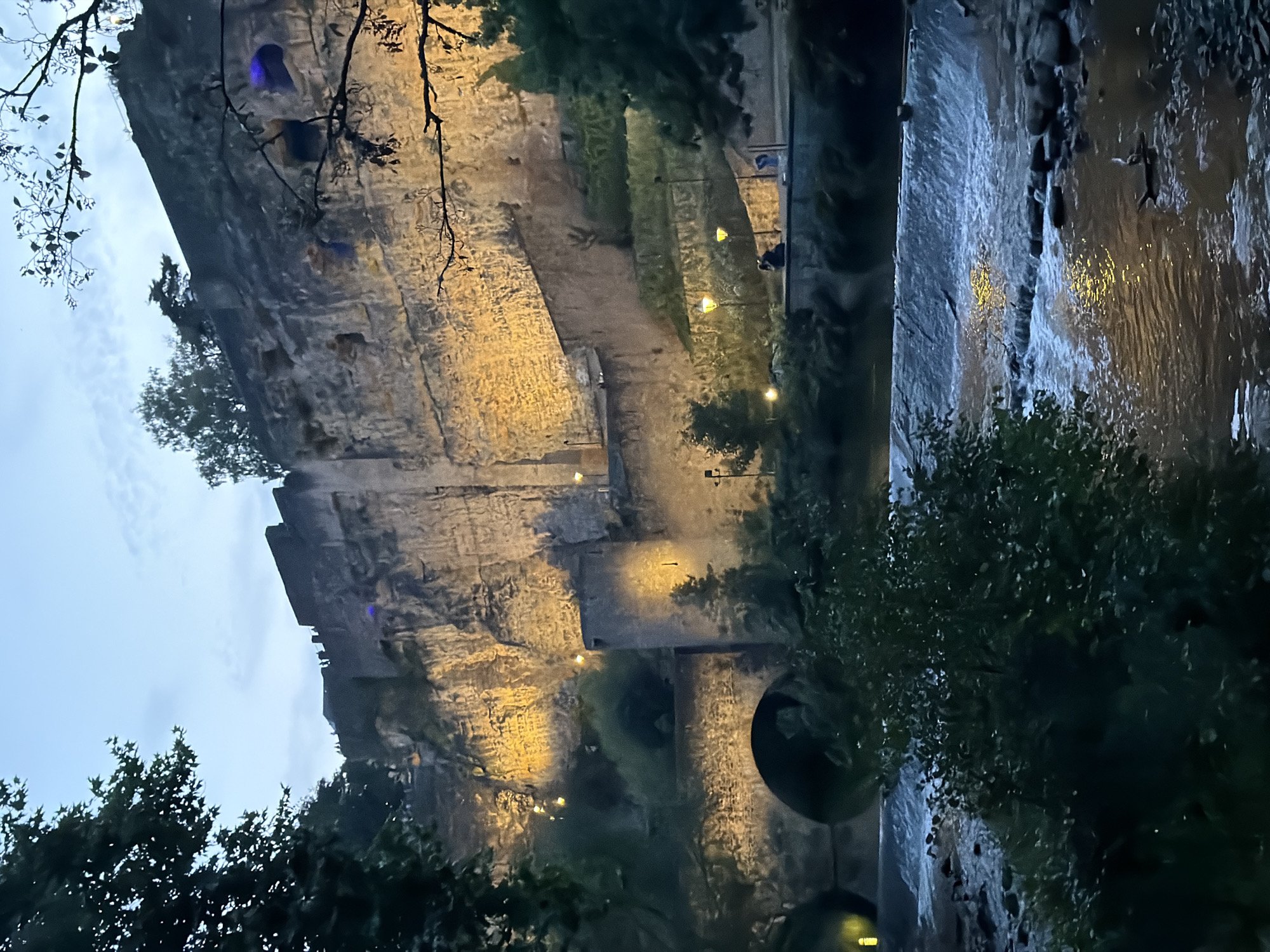 The image shows the Bock Casemates in Luxembourg City, Luxembourg, at dusk. The imposing stone fortifications, illuminated by warm yellow lights, rise from a steep cliff face. A river or canal flows in the foreground, with a small waterfall or weir creating ripples and whitewater. Trees and foliage frame the scene, with some branches silhouetted against the twilight sky. Several figures are visible on a pathway along the base of the fortifications, suggesting the location is accessible to visitors. The overall atmosphere is serene and historic, with the artificial lighting highlighting the ancient architecture against the fading natural light.