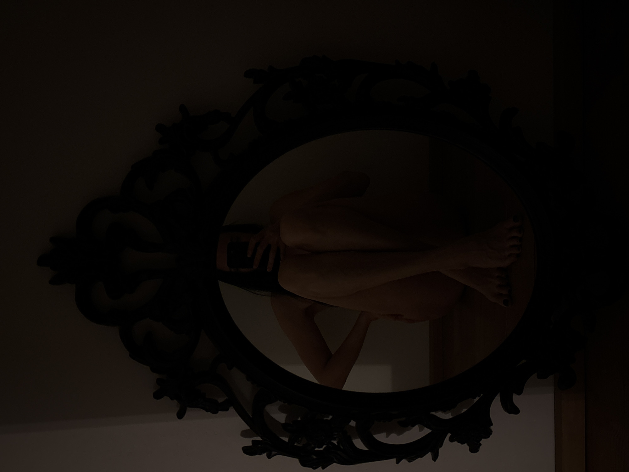 The image depicts a person reflected in an ornate, oval-shaped mirror with a dark, possibly black, frame. The person is positioned within the mirror's reflection, appearing to be seated with their legs drawn up towards their body and arms wrapped around their legs. One hand seems to be holding a mobile phone. The setting is dimly lit, with the wall behind the mirror a light color. The reflection is upside down relative to the mirror frame, suggesting an intentional artistic composition. The location in Chaikovsky, Russia is unknown.