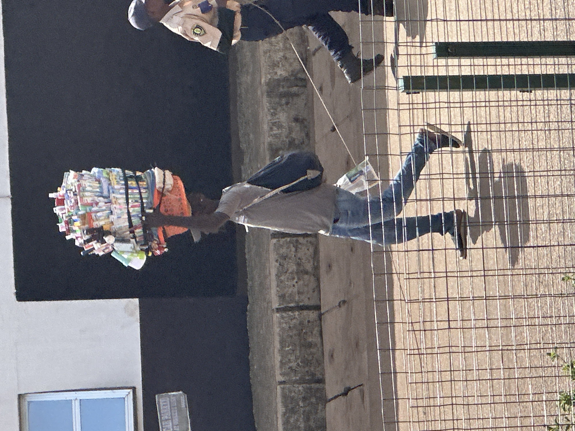The image shows two people in what appears to be a market or street setting in Bukuru, Nigeria. One person is wearing a light-colored uniform with a patch on the left chest and a dark cap. They are standing behind a large bundle of items strapped together with black bands. The bundle appears to be made up of many small, colorful packages, possibly toiletries or other small goods.
The second person is a vendor, also likely in a market setting. They are carrying a large, colorful bundle of items on their head. These items are also strapped together and appear to be a variety of small, individually wrapped products. The vendor is wearing a light-colored t-shirt and jeans, and is walking towards the right side of the frame. They are walking behind a metal fence or barrier.
The scene is outdoors, with sunlight casting shadows, suggesting it is daytime. The background includes a dark wall on the left and a textured, light-colored wall on the right, with a metal fence structure. The overall atmosphere appears to be busy, characteristic of a marketplace. There is no visible text in the image that is legible.