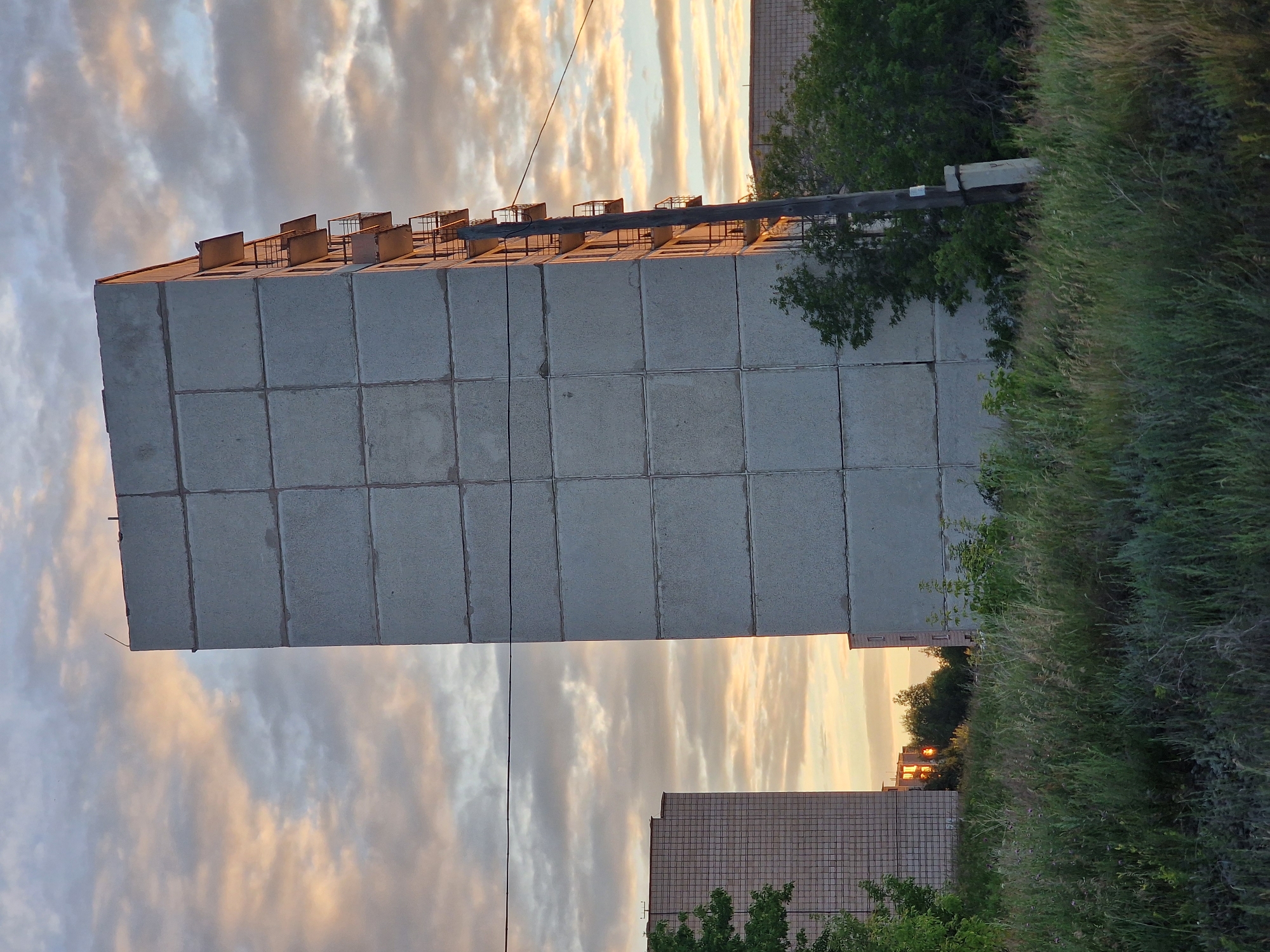 An image captures a concrete apartment building in Kashgar City, China, during what appears to be sunset. The sky is filled with a mix of clouds, some tinged with orange and yellow from the setting sun, while others are a pale blue and gray. The building itself is tall and rectangular, constructed from large, square concrete panels. Balconies with rusty metal railings jut out from one side of the building, spaced evenly. A weathered wooden utility pole stands near the building, supporting a single black wire that stretches across the frame. In the foreground, tall, dry grass and weeds create a wild, overgrown field. To the left, another apartment building with a grid-like facade is visible, and to the right, more trees and another building can be seen in the distance. The overall scene is somewhat stark, with the industrial architecture contrasted by the natural elements and the soft, atmospheric light of the sky. There are no people visible in the image.