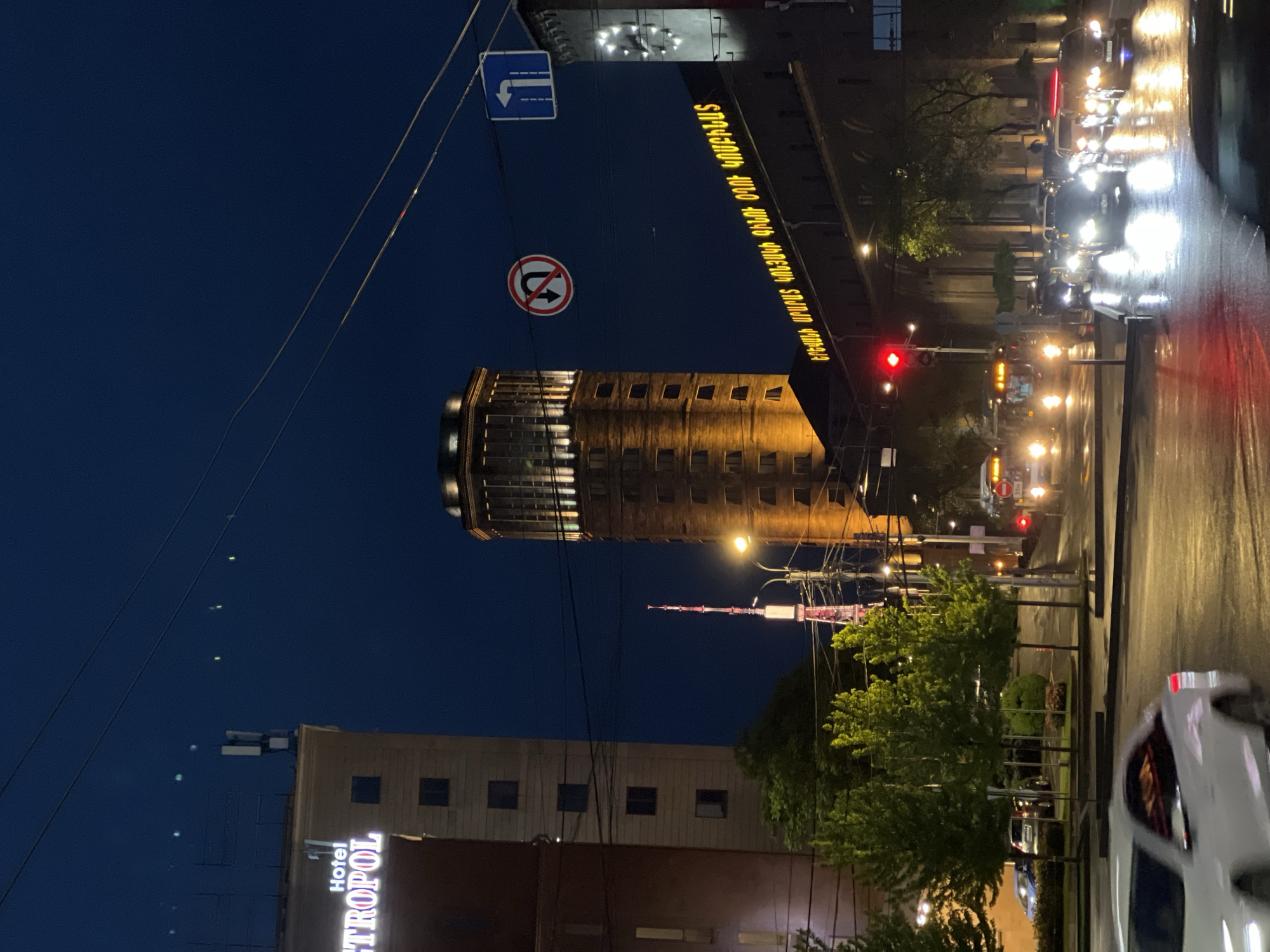 The image presents a nighttime urban scene in Yerevan, Armenia. Prominently featured is a cylindrical brick tower, likely a historical or architectural landmark, illuminated by warm artificial light. Overhead electrical wires crisscross the sky, partially obscuring the view of the tower.
Streetlights line the roadway, which reflects the lights from passing vehicles, creating streaks of white and red. A digital display with text in Armenian is visible above a building or structure.
In the lower portion of the image, the "Hotel Metropol" sign is partially visible. Also, a television tower with red and white segments is visible further behind the cylindrical tower. Street signs are visible above the cylindrical tower.
The image is taken from a low angle, possibly from street level. The overall environment suggests a busy city street with a mix of historic architecture and modern infrastructure.