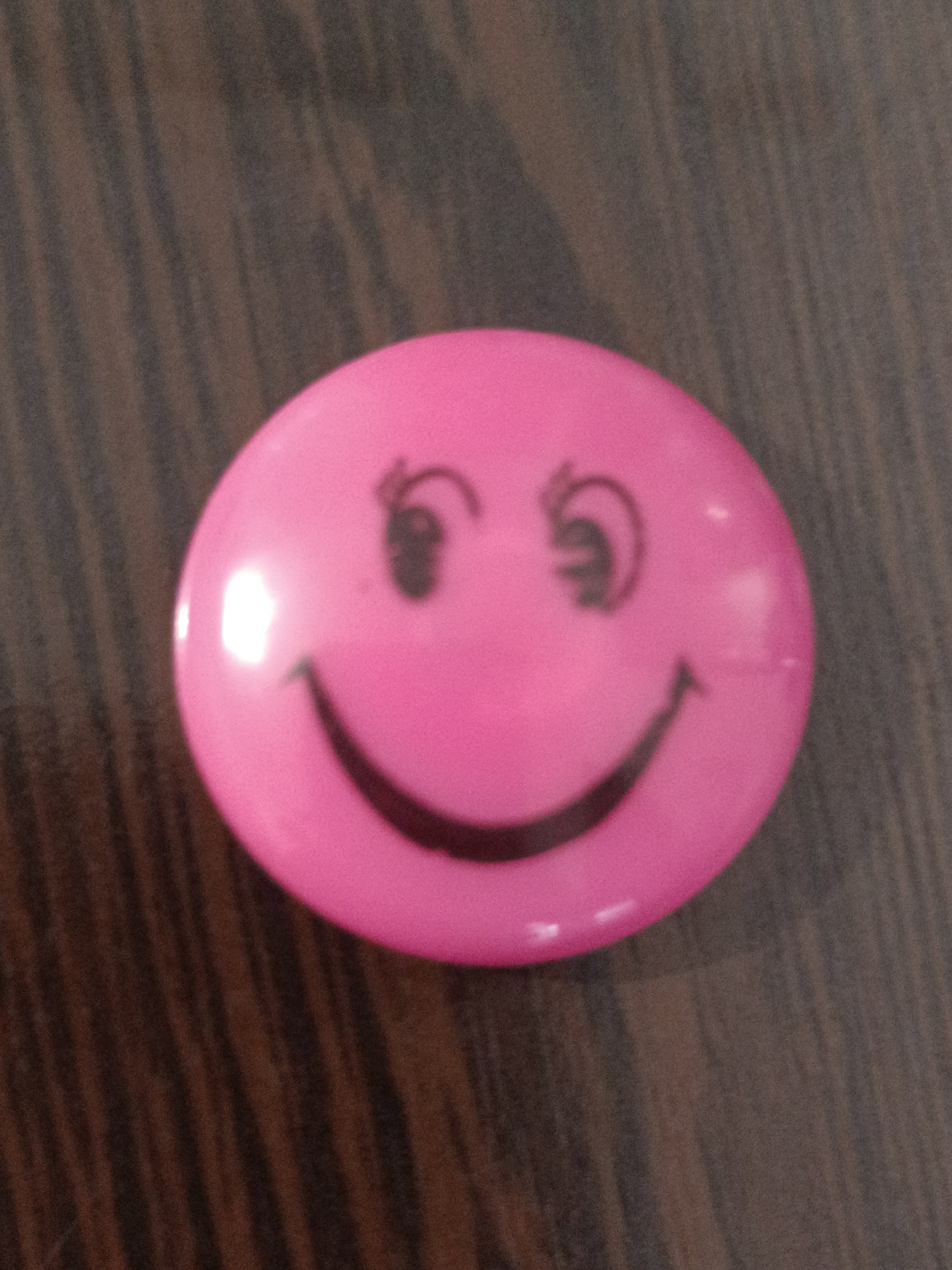 A close-up, slightly blurry, overhead shot features a pink, oval-shaped object with a smiley face drawn on it. The object is placed on a dark, wooden surface with a visible wood grain pattern. The smiley face is depicted with two simple, dark eyes and a wide, curved smile. The background is out of focus, with the wooden surface filling the majority of the frame. The lighting appears to be from above, causing a bright highlight on the left side of the pink object. The location in the city of Taraz city administration, Kazakhstan is not discernible from the image.