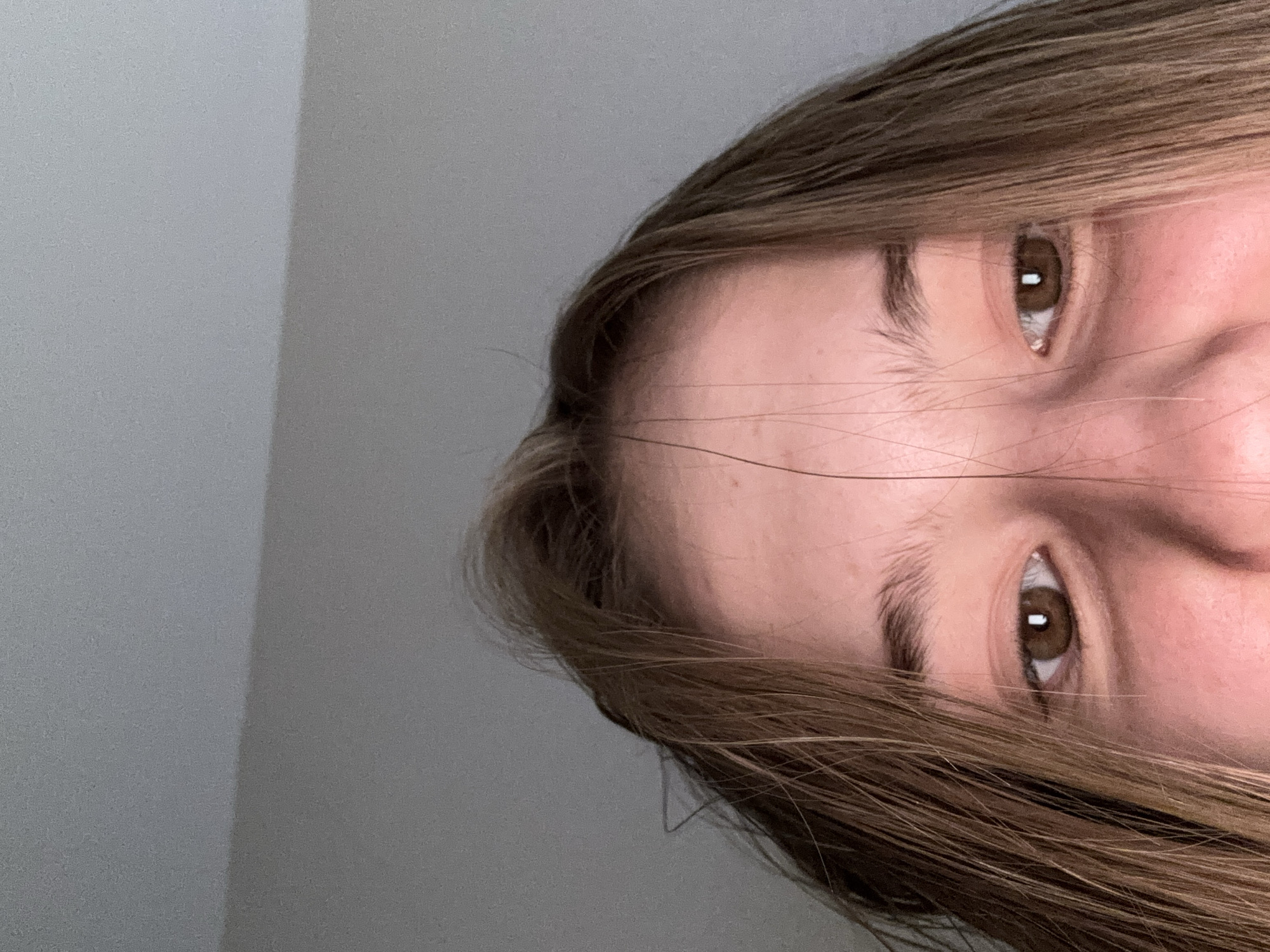 The image is a close-up, overhead shot of a young person's face, focusing on their eyes and forehead. The person has light brown hair that falls in strands around their face. Their eyes are open and looking forward, with brown irises. The skin on their face appears slightly flushed, and the background is a plain, grey wall. The image does not contain any elements that would allow for the determination of a specific city, country, or location, including Sinyavino, Russia.