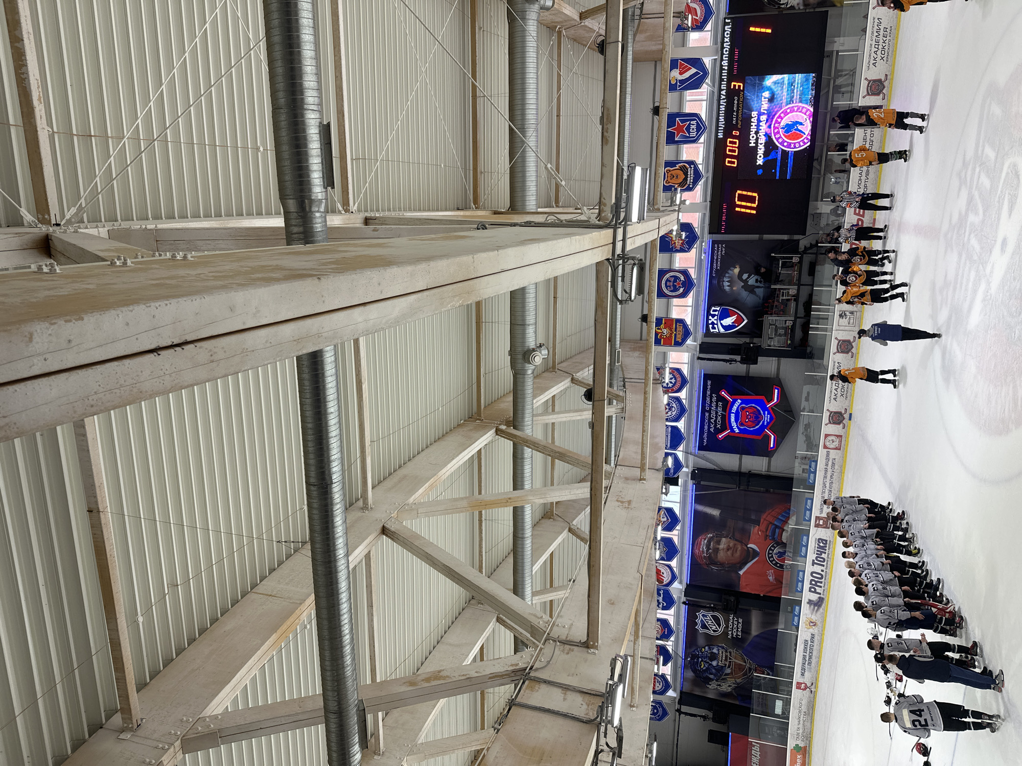 The image shows the interior of a hockey arena in Chaikovsky, Russia. A large scoreboard displays the score as 10-3. Hockey teams are lined up on the ice. Banners displaying sports teams and logos hang on the wall. The arena's ceiling features exposed structural beams and ductwork.