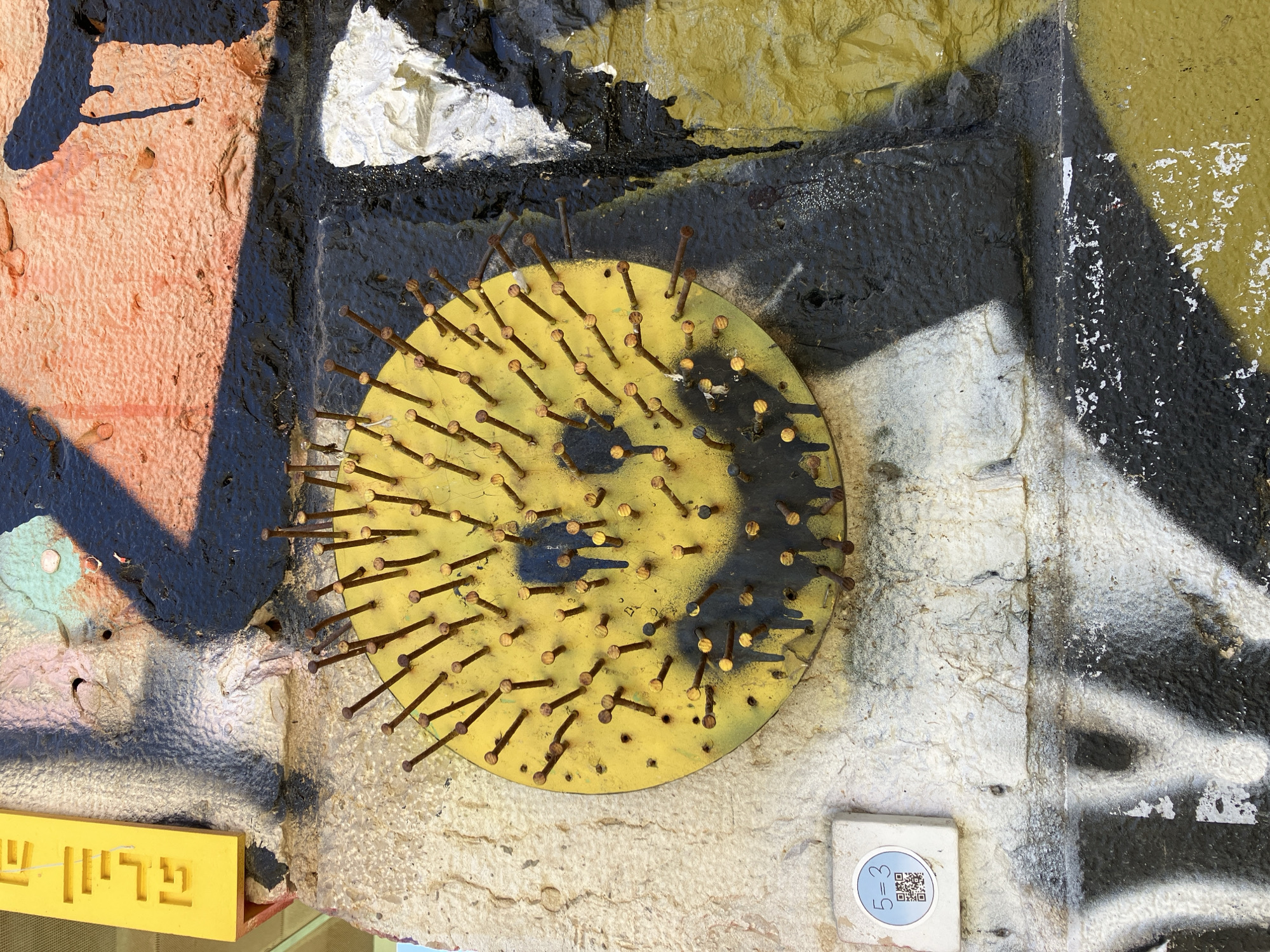 The image shows a yellow, circular metal plate affixed to a textured wall covered in graffiti. The plate is studded with numerous rusty nails protruding outwards, arranged to partially form a smiley face with dark paint. The surrounding graffiti includes a mix of colors and textures, including peach, black, white, and yellow paint on rough surfaces. A small, square white tile with a QR code and some text is located below the metal plate. Part of a yellow sign with Hebrew writing is visible in the lower left corner. The location is likely in Tel-Aviv, Israel, as indicated by the Hebrew writing.