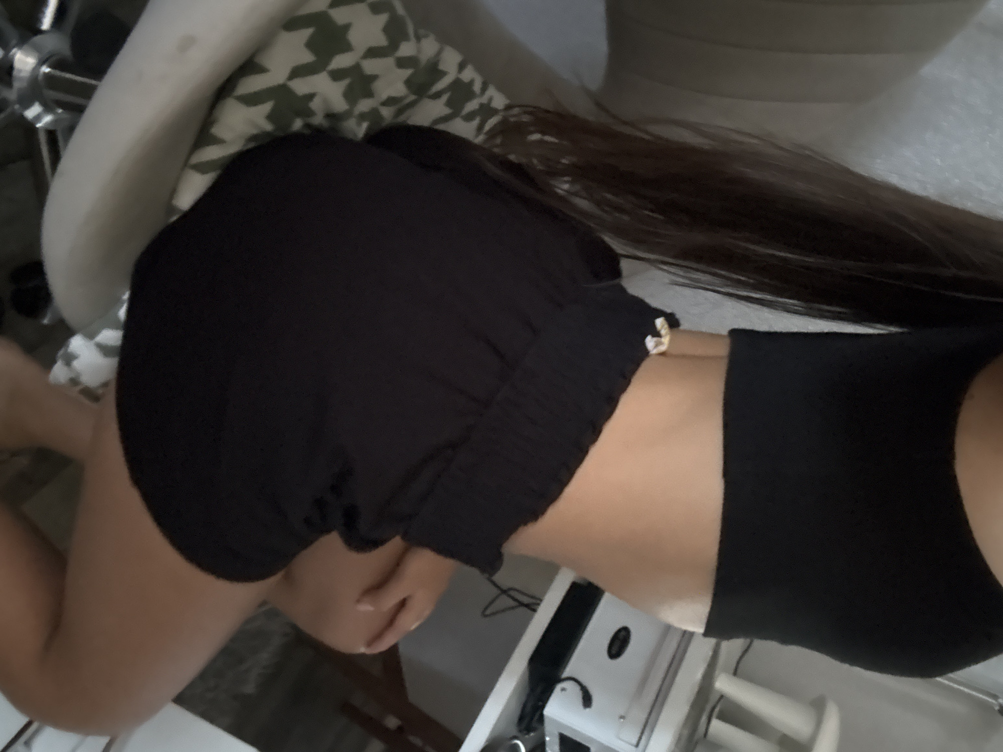 A person, viewed from the side and partially from the back, is seated in a chair. They are wearing a black crop top and black shorts. Their long, dark hair is visible, cascading down their back. The person's skin tone is light. The background shows a desk with a white appliance on it, and part of a patterned cushion on the chair. The environment appears to be indoors, possibly a room in a house or office, but the specific location in Basarabeasca, Moldova, cannot be determined from the visual information.
