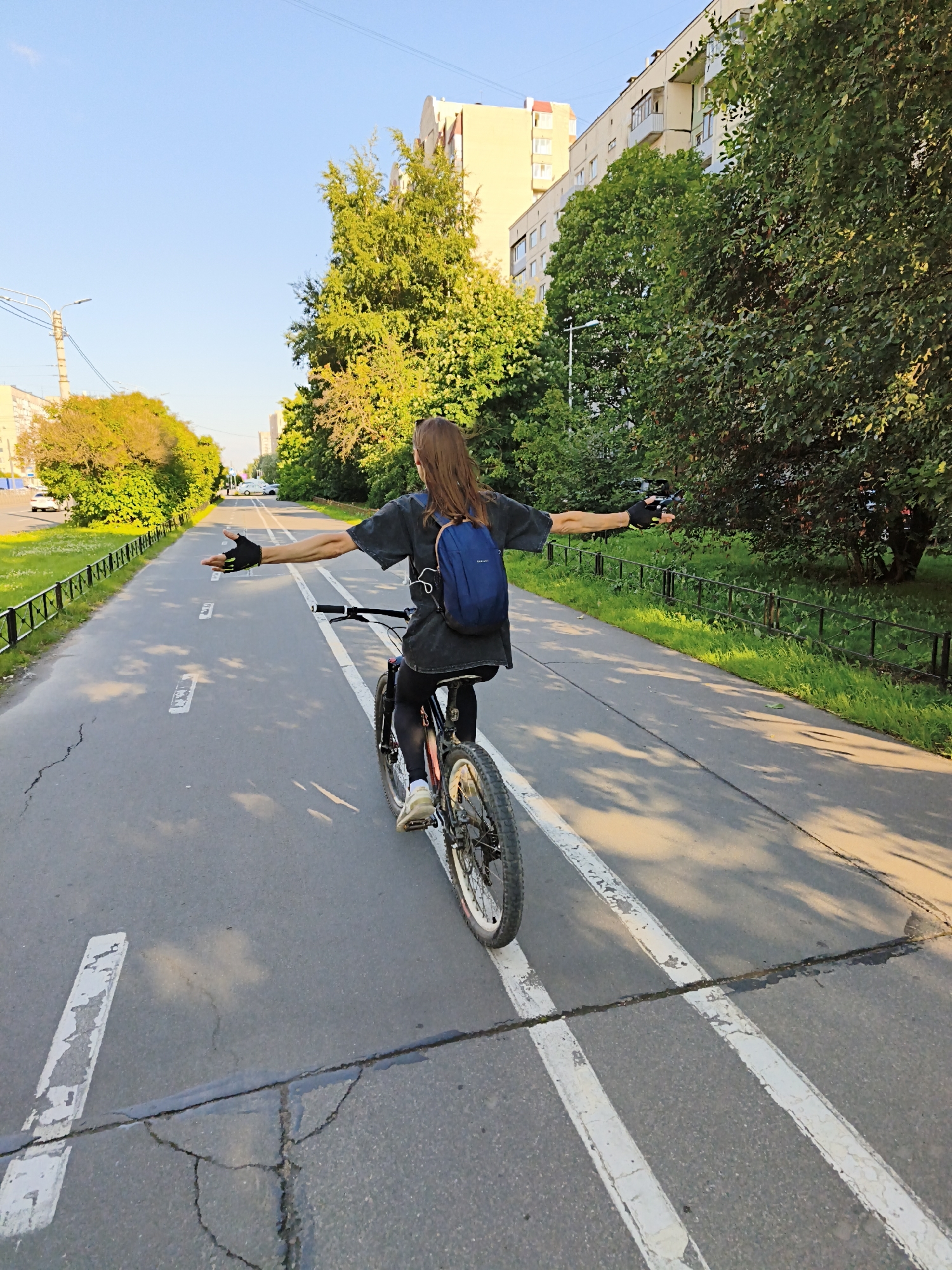 A person is riding a bicycle on a paved path with white lane markings. The person is facing away from the camera, with their arms extended outwards. They are wearing a dark t-shirt, black leggings, and a blue backpack. The bicycle has a dark frame and spoked wheels. The path appears to be in an urban environment, with buildings and trees visible in the background. The sky is clear and blue, and the sun is casting shadows on the ground. The overall scene depicts a person enjoying a bicycle ride in a city park or on a dedicated bike path.