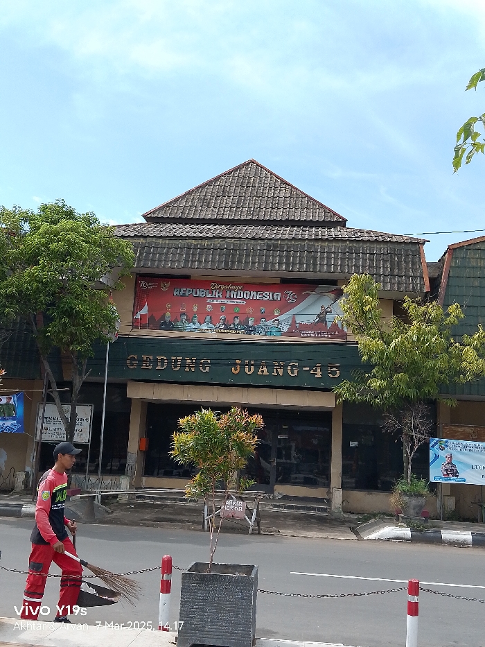 The image shows a street scene in Indramayu, Indonesia, located at coordinates -6.326, 108.323. A single person, a man wearing a red and white shirt, is sweeping a sidewalk in front of a building. The building has a tiled roof and a sign that reads "GEDUNG JUANG-45." Above the sign is a banner with the text "Dirgahayu Republik Indonesia 78" and images of Indonesian leaders. There are two trees and a planter box in the foreground, and a chain-link fence lines the street. The sky is blue and clear, and the weather appears to be sunny. A red "Awas Motor" sign is visible near the planter. A sign that reads "Pembinaan Satuan" is visible near the building. There is a large banner on the right side of the building that features an image of a man with a motorcycle helmet. The image was taken at 14:17 on March 7, 2023.