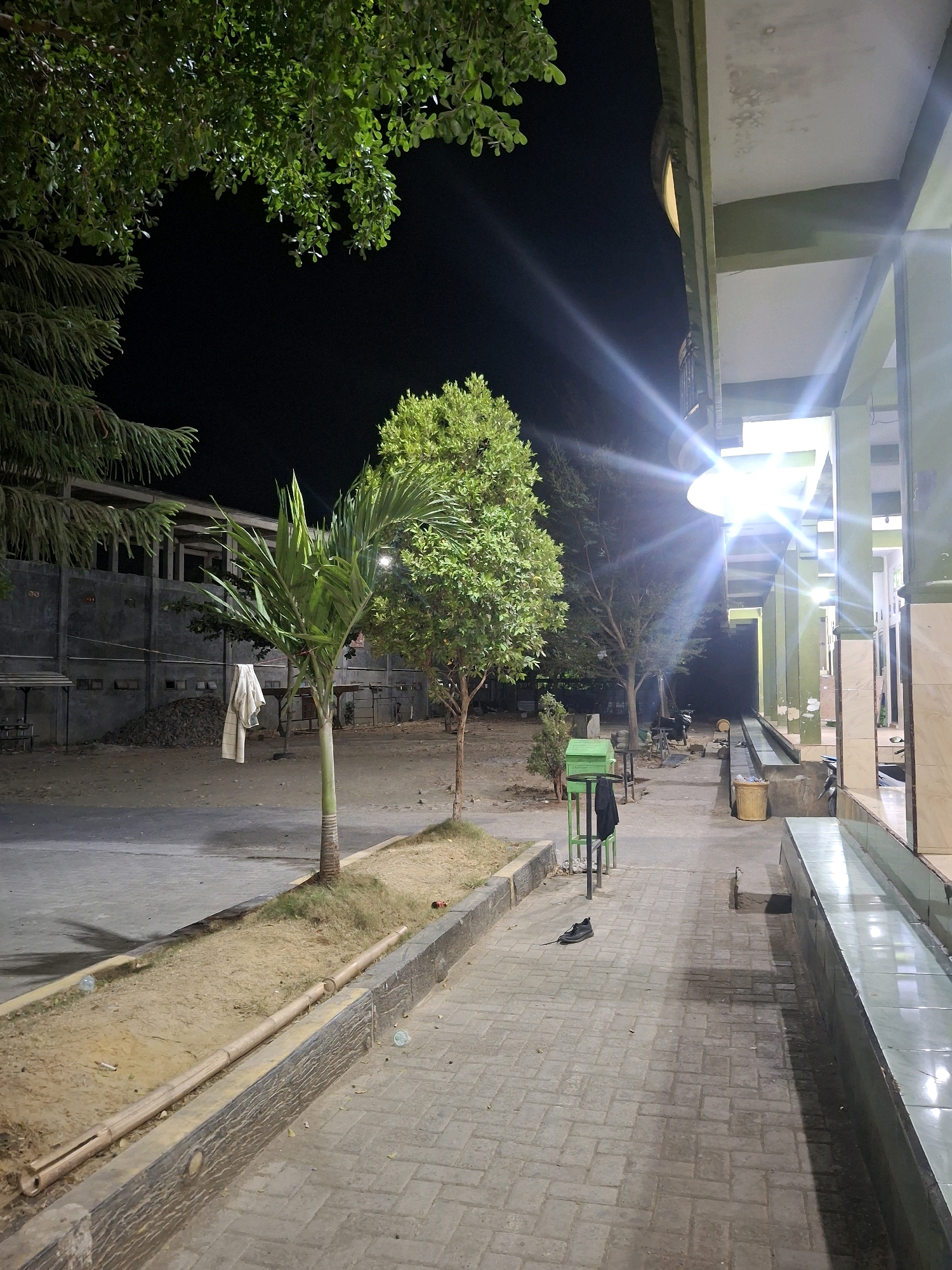 The image shows an outdoor area at night, likely in Potoan Laok, Indonesia, adjacent to a building with an extended covered walkway. A bright light shines from the building, creating a strong glare. The ground is paved with rectangular bricks, and a raised, sandy planter with bamboo edging runs along the side. Several trees are present, including a young palm tree and other leafy trees. A white garment hangs from a clothesline strung between two poles in the background. A green metal trash receptacle with a black garment draped over it stands near the walkway. A single black shoe rests on the brick pavement. In the distance, a wall with visible concrete structures and piles of rubble is visible. The scene appears to be a public space, possibly near a mosque or community center, with an emphasis on nighttime lighting and an outdoor area that has been maintained.