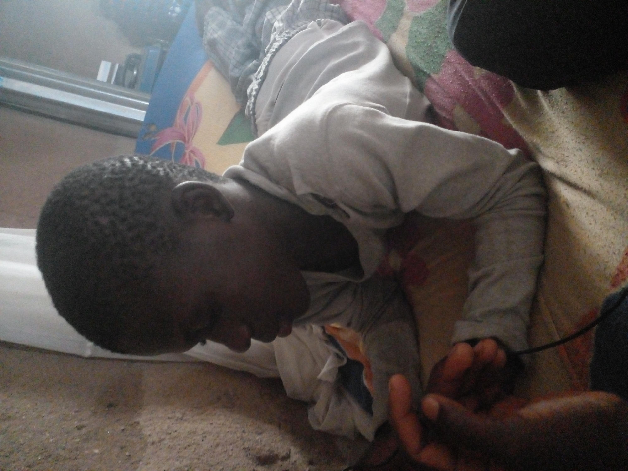 This image shows a person lying on their side on a bed. The person is wearing a light gray long-sleeved shirt and light-colored pants. Their head is resting on a pillow, and their face is partially visible, looking downwards. The person's right hand is reaching out and appears to be holding another hand, which is out of focus and darker in color.
The bed is covered with a patterned blanket that has shades of pink, green, and white. A blue object with a pink floral design is visible behind the person. The background is somewhat blurry but suggests an indoor setting. The lighting appears to be natural, possibly from a window, casting shadows and highlights on the subjects and the bedding. The overall mood seems calm and intimate, given the gentle gesture of holding hands. The image quality is moderately low, with some graininess and softness. There is no visible text in the image.
