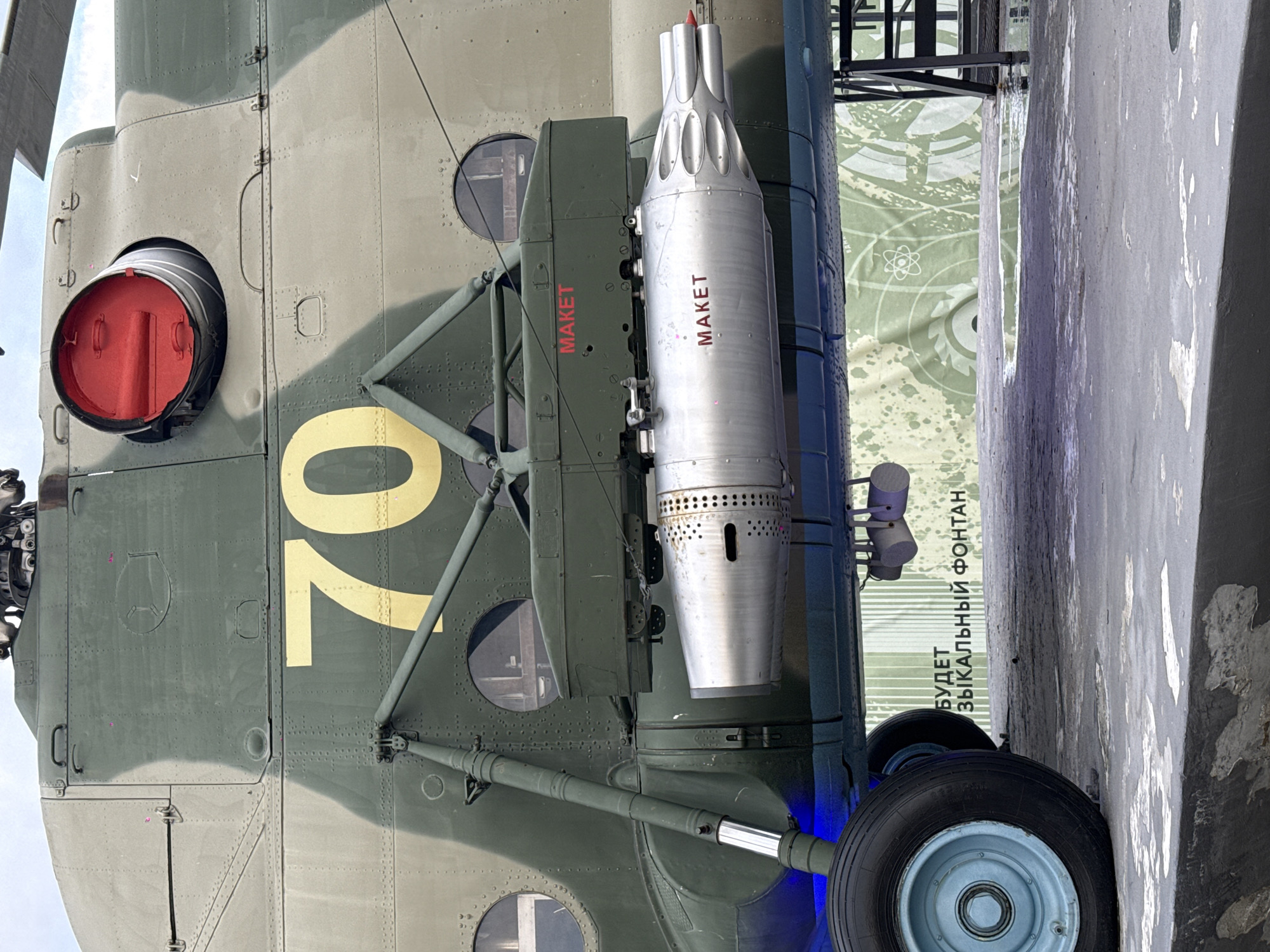 The image shows the side of a camouflaged helicopter with the number "70" painted on it in gold. A silver pod-like object with "MAKET" (model) written on it in red is attached to the side of the helicopter. The landing gear and wheel are also visible. The text "БУДЕТ ЗЫКАЛЬНЫЙ ФОНТАН" (There will be a musical fountain) is visible on a nearby structure. The image was taken in Moscow, Russia.