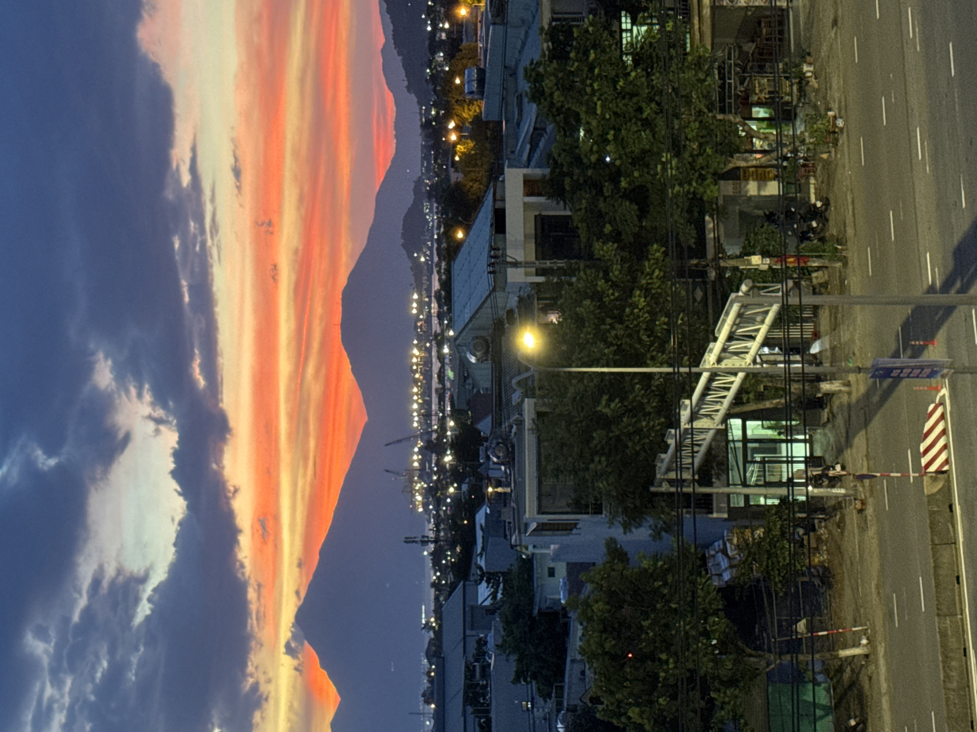 The image depicts a panoramic view of a city at dusk, likely in Đà Nẵng, Vietnam, given the distinctive coastal landscape with mountains in the background and the urban sprawl in the foreground.
The sky is a dramatic canvas of colors, transitioning from deep blues and purples at the top to vibrant oranges and yellows near the horizon. The vibrant hues suggest a sunset or sunrise, casting a warm glow over the scene. Fluffy clouds are scattered across the sky, adding texture and depth to the atmospheric conditions.
In the distance, silhouetted against the colorful sky, are the majestic forms of mountains. Beyond the mountains, a body of water is visible, reflecting the twilight colors. A port or industrial area is situated along the coastline, marked by the presence of cranes and numerous bright lights, indicating activity even as daylight fades.
The mid-ground of the image reveals the urban landscape of Đà Nẵng. Buildings of various sizes and architectural styles are clustered together, some appearing to be residential, while others might be commercial establishments. The rooftops are predominantly tiled, a common feature in Vietnamese architecture. Trees are interspersed throughout the urban environment, their dark foliage contrasting with the illuminated buildings.
The foreground shows a street scene. A paved road with white lane markings stretches across the bottom of the frame. A concrete barrier separates the road from a sidewalk or pedestrian area. On the sidewalk, a pedestrian overpass with a white metal structure is visible, spanning across the road. Streetlights illuminate sections of the scene, casting pools of yellow light. Several parked motorcycles are visible, suggesting typical urban transportation. A traffic sign with red and white markings is positioned near the overpass, and a traffic cone is placed on the road. The overall impression is a bustling city transitioning into night, with the natural beauty of the landscape providing a stunning backdrop.