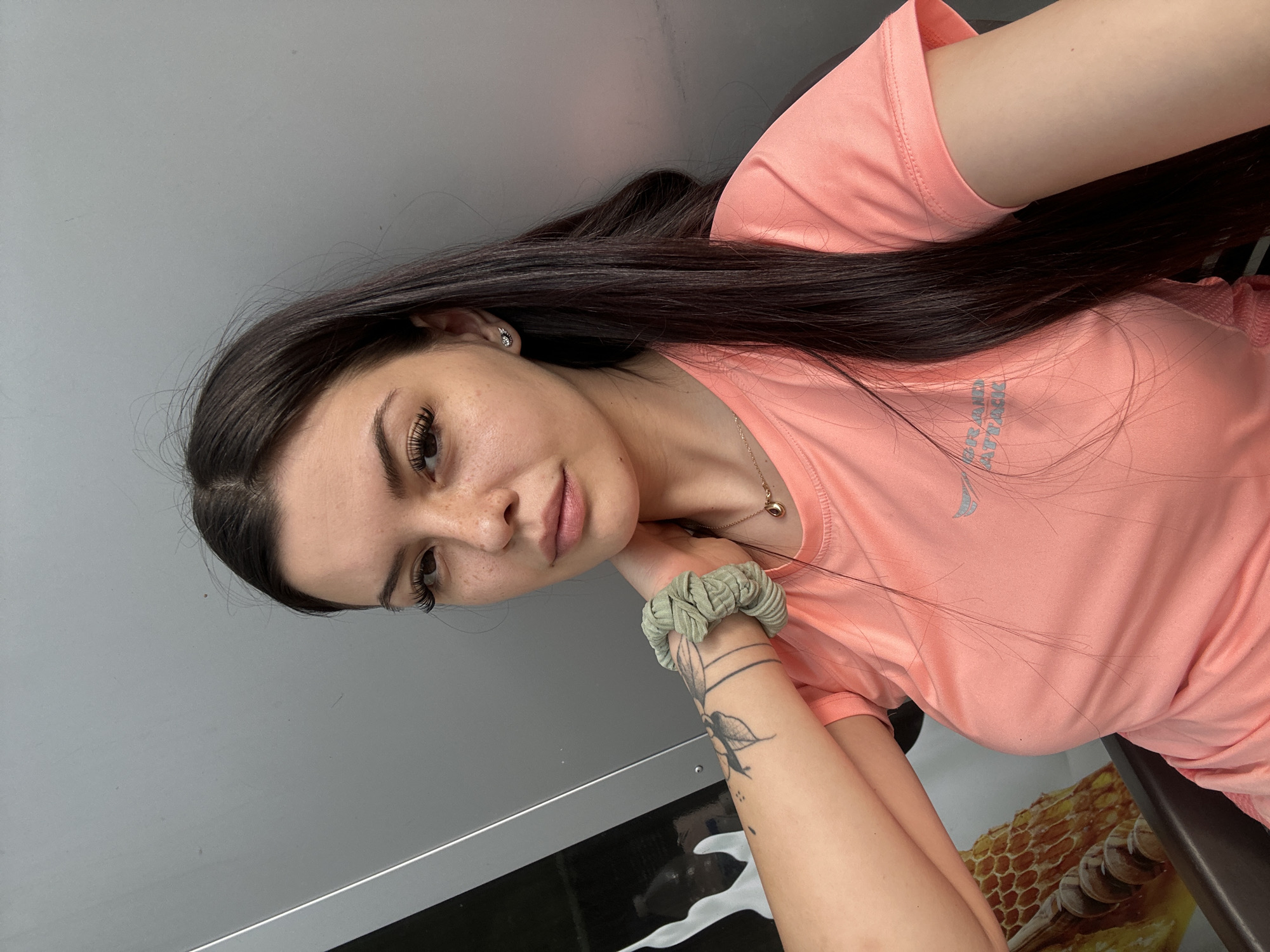 A young woman with long, dark hair and fair skin is looking directly at the camera. She has noticeable freckles on her face and is wearing a peach-colored t-shirt. A scrunchie is on her left wrist, and a tattoo is visible on her left forearm. She is also wearing a delicate necklace with a pendant and small wing-shaped earrings. The background appears to be a textured, gray surface, possibly a wall or a vehicle. The image was taken in Sliven, Bulgaria.