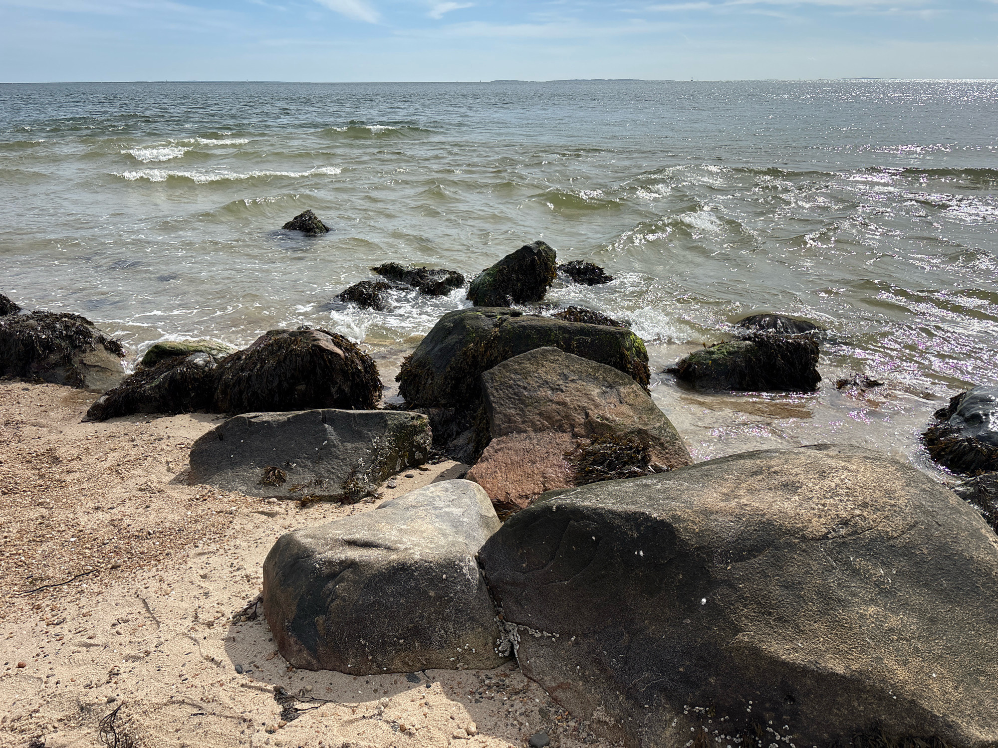 The image depicts a rocky beach scene with the ocean in the background, likely located in Waterford, United States of America. The foreground features a sandy beach littered with various sized rocks, some covered in seaweed. The water is choppy with small waves lapping at the shore. The sky is partly cloudy. No people or animals are visible in the frame. The overall impression is one of a natural, coastal environment.