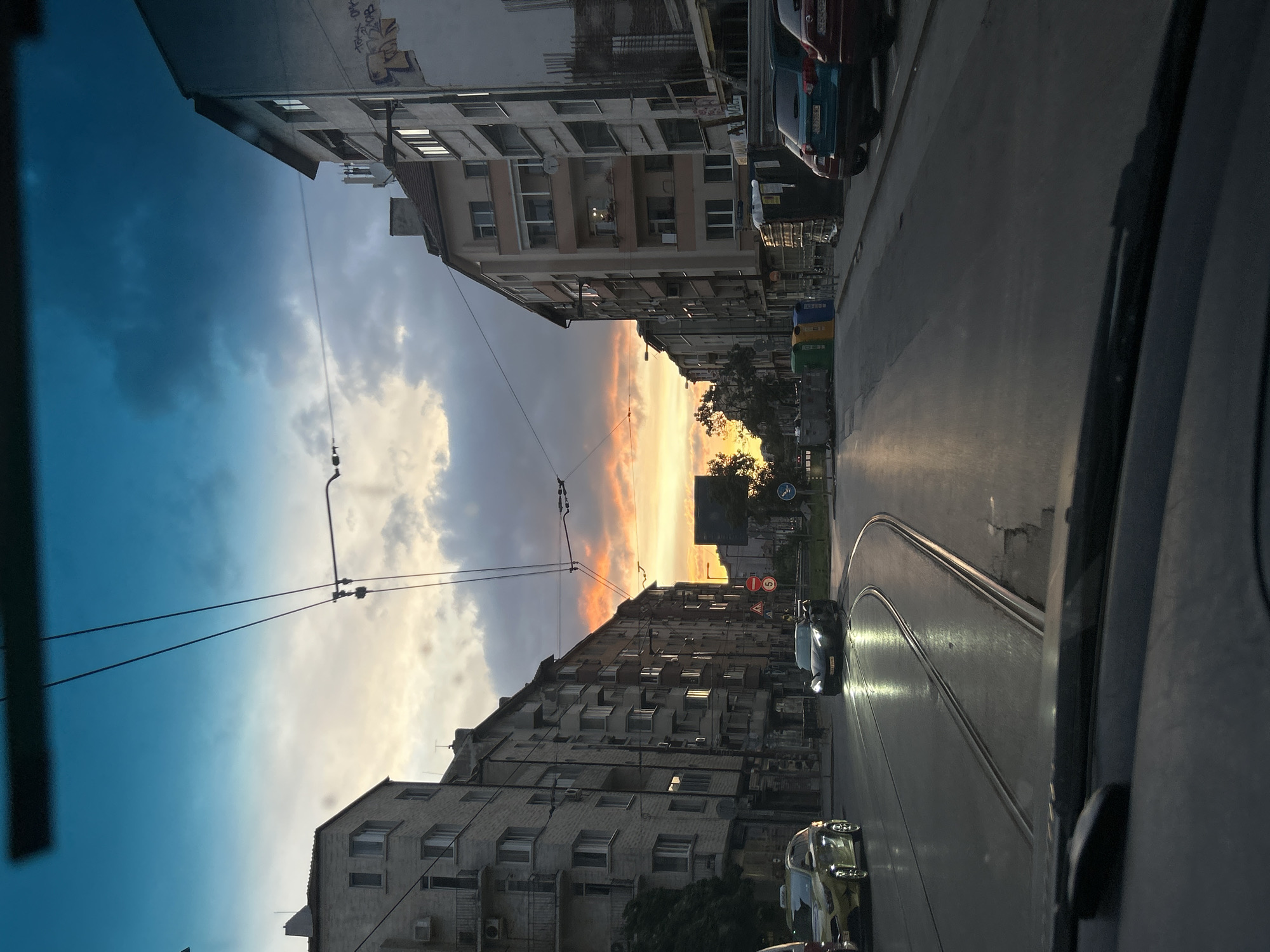 The image captures a street scene at sunset in an urban environment, likely a city in Eastern Europe due to the architecture and tram lines. The viewpoint is from inside a vehicle, looking forward through the windshield. The sky is dramatic, with hues of blue, grey, and orange from the setting sun, illuminating the clouds.
On the left side of the street, a row of apartment buildings with balconies and visible windows stretches into the distance. The buildings appear to be aged, with some showing signs of wear. A yellow car, possibly a taxi, is parked on the left.
The street itself is paved and features tram tracks that curve towards the center of the image. A dark-colored sports car is driving on the street, its headlights illuminated and reflecting on the wet asphalt.
On the right side of the street, another row of apartment buildings is visible, some with graffiti on the walls. Several parked cars are visible, including a teal SUV and a red car. Large blue and green dumpsters are also present on the right side of the road.
Overhead, tram wires crisscross the sky, supported by poles, suggesting an active public transportation system. A traffic sign is visible in the middle distance, and trees are scattered along the sides of the street. The overall impression is a typical city street during dusk, with a mix of residential buildings, traffic, and infrastructure.