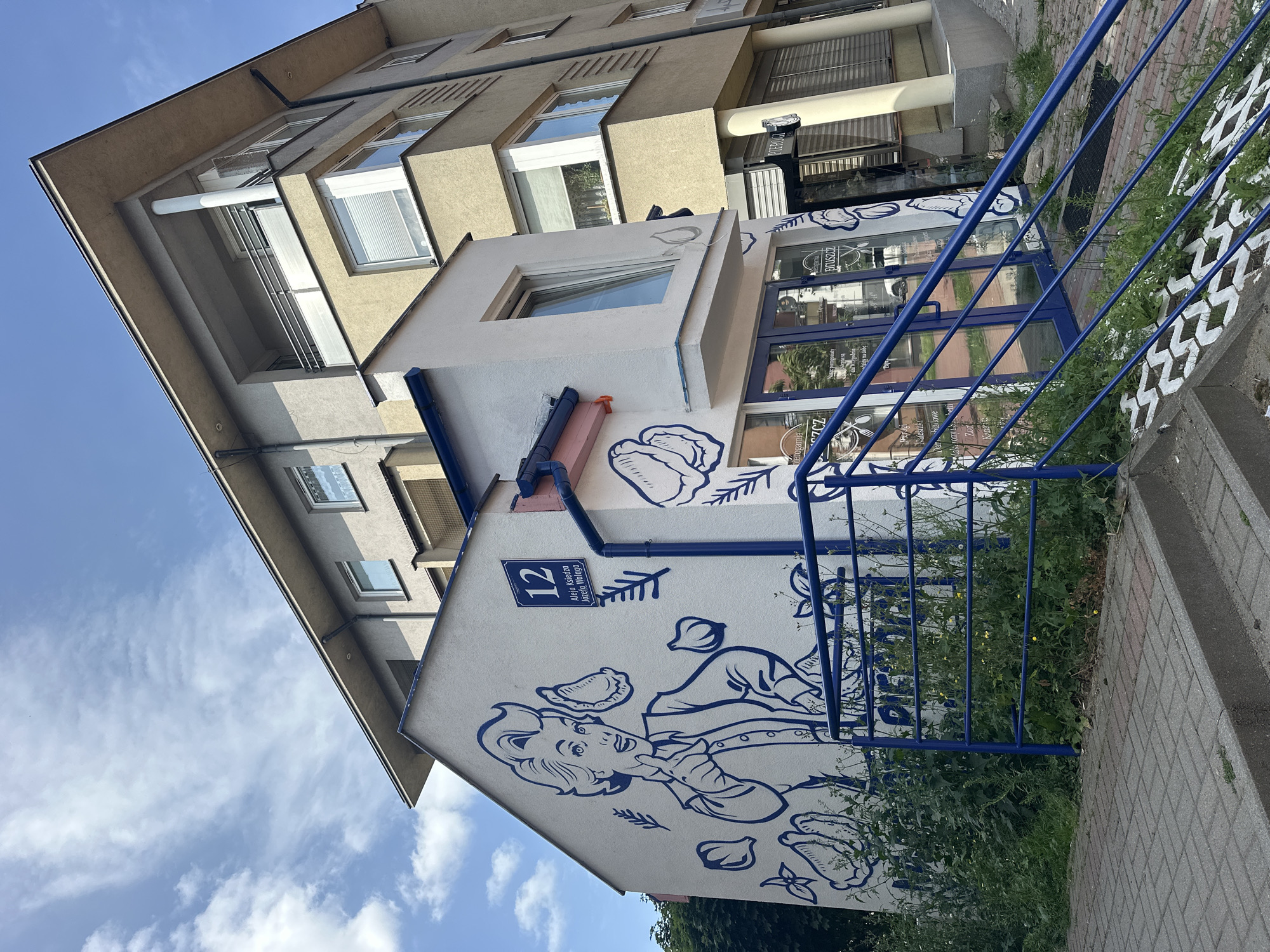 This is a medium-shot, eye-level, outdoor photograph taken on a sunny day in Pruszcz Gdański, Poland. The image features a two-story building with a mural on its side. The building has a white facade with blue accents and a blue railing leading up to its entrance. The mural depicts a cartoonish woman pointing to the right, with images of food items such as garlic and dumplings surrounding her. A blue sign with the number "12" and Polish text "Aleja Księdza Józefa Waląga" is affixed to the side of the building, indicating the street address. The building is situated next to a larger, multi-story apartment building with a beige facade. In the foreground, there are paved steps and an overgrown grassy area. The sky above is blue with scattered white clouds. The overall scene suggests a commercial establishment, likely a restaurant or food shop, decorated with a vibrant mural.