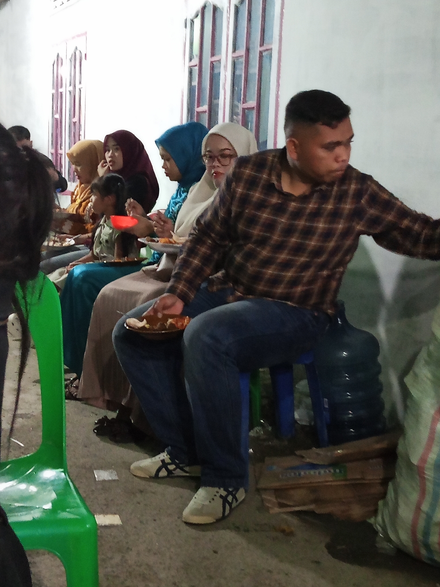 A group of people are sitting on the ground and a chair, appearing to be at a gathering or meal in Perbarakan, Indonesia. The individuals, some wearing hijabs, are holding plates of food. A man in a plaid shirt is seated on a blue stool next to a large water jug. A bright green plastic chair is positioned to the left of the frame, and a large, striped bag is visible on the right. The setting seems to be outdoors, possibly a residential area, with a white wall and windows in the background.