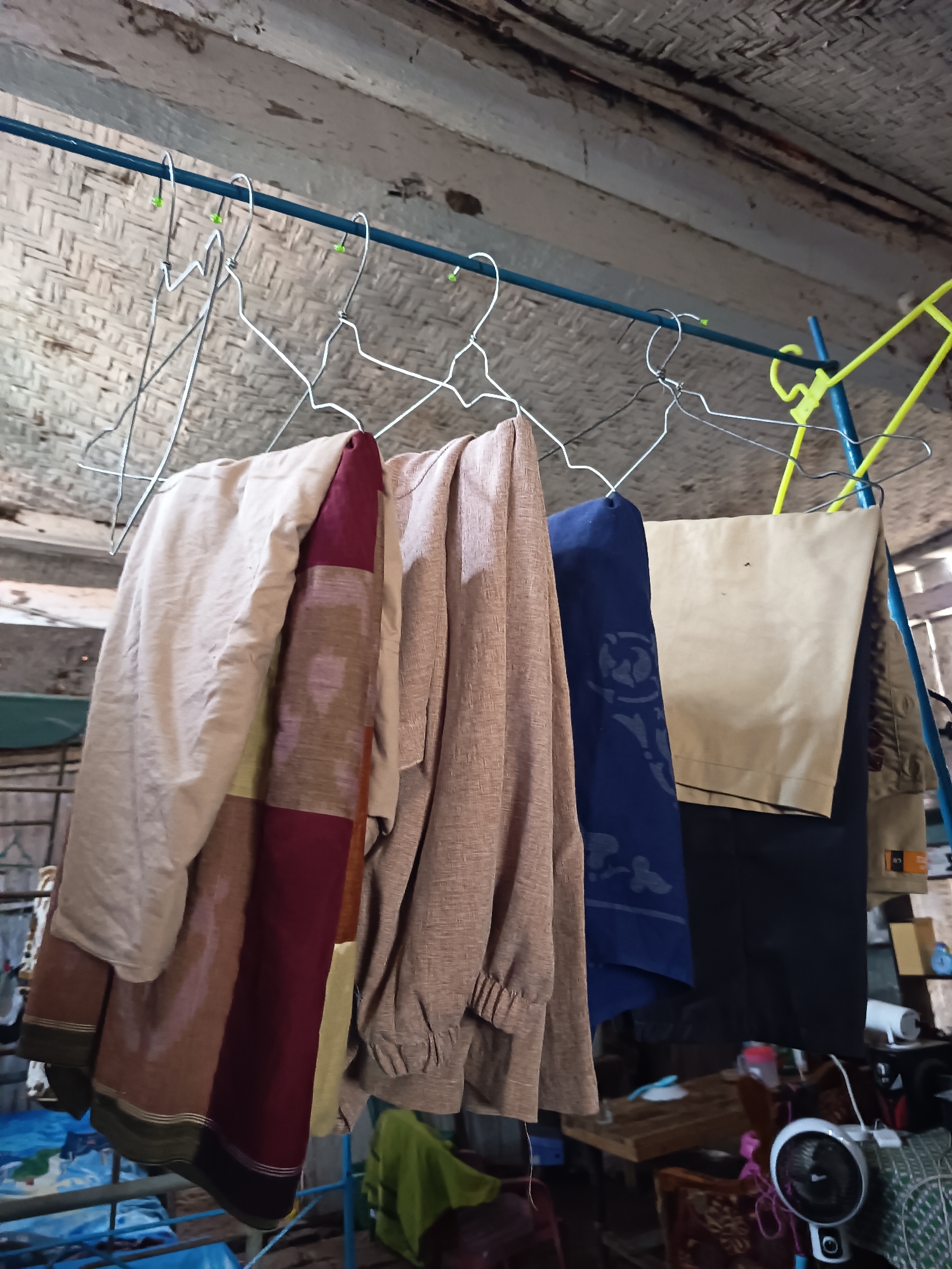 In Krampilan, Indonesia, an indoor drying rack displays several garments. The rack, made of a blue metal pipe supported by metal poles, is suspended from the ceiling. Several wire hangers hold clothing items: a light-colored piece of fabric, a garment with burgundy, brown, and cream colored stripes, a brown textured fabric, and a navy blue garment with a patterned design. To the right, a tan colored fabric hangs, partially obscured by a black garment. The background consists of a textured wall and ceiling made of woven material, suggesting a rustic interior. Various objects are visible on shelves and tables below the drying rack, including a small white fan, containers, and what appears to be a blue striped mattress.