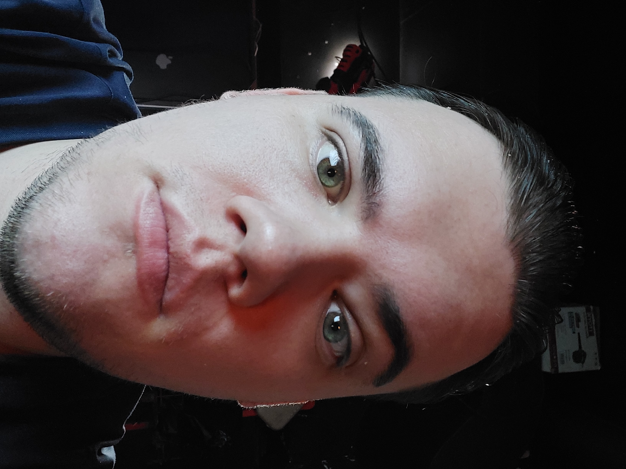 A close-up, slightly rotated portrait of a light-skinned male. The subject has short, dark hair that appears slightly wet. He has light green eyes and a faint stubble beard. He has a slight smirk or subtle smile. Behind him are indistinct shapes and objects in dark colors, including what appears to be an Apple laptop (as indicated by the logo). The background is dark, obscuring any clear details about the environment. There's a red and black object behind the subject's head as well as what appears to be a cardboard box. Location details (Tashkent, Uzbekistan) are not discernible from the image provided.