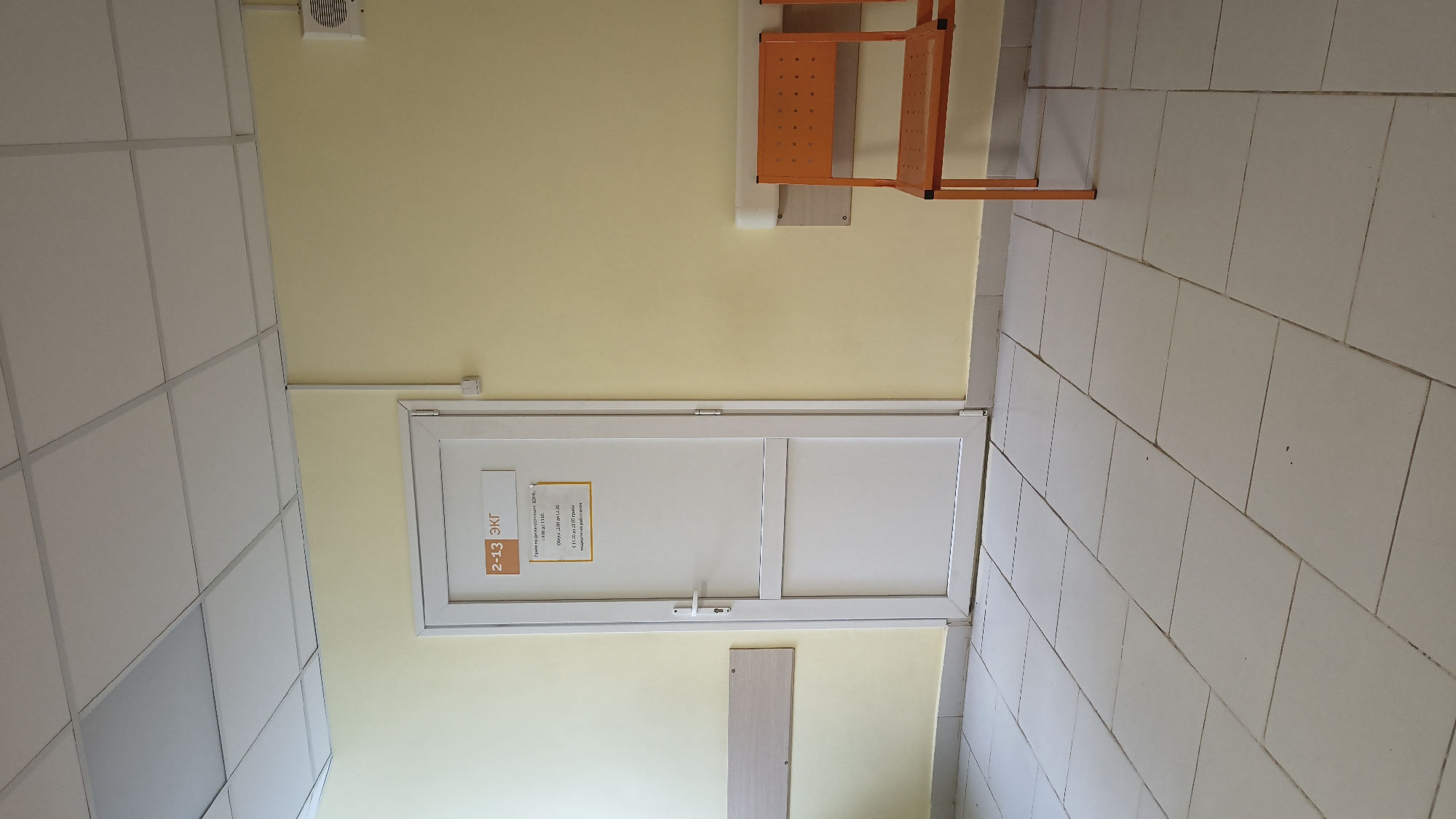 The image shows a hallway in a medical facility. On the left, a white door with a sign that reads "2-13 ЭКГ" (2-13 ECG) is visible. Below the room number, there is a schedule indicating the operating hours for ECG services: "Утреннее время (запись): с 8:00 до 14:00" (Morning hours (appointment): from 8:00 to 14:00) and "Дневное время: с 14:00 до 19:00" (Afternoon hours: from 14:00 to 19:00), and "Ежедневно с 8:00 до 20:00" (Daily from 8:00 to 20:00). A light beige wooden panel is mounted on the wall to the left of the door. On the right side of the hallway, two orange chairs are positioned against a pale yellow wall. A white ventilation grille is visible on the upper right portion of the wall. The floor is tiled with light gray square tiles. The lighting appears to be from overhead fluorescent lights. The overall scene suggests a waiting area or corridor within a clinic or hospital.