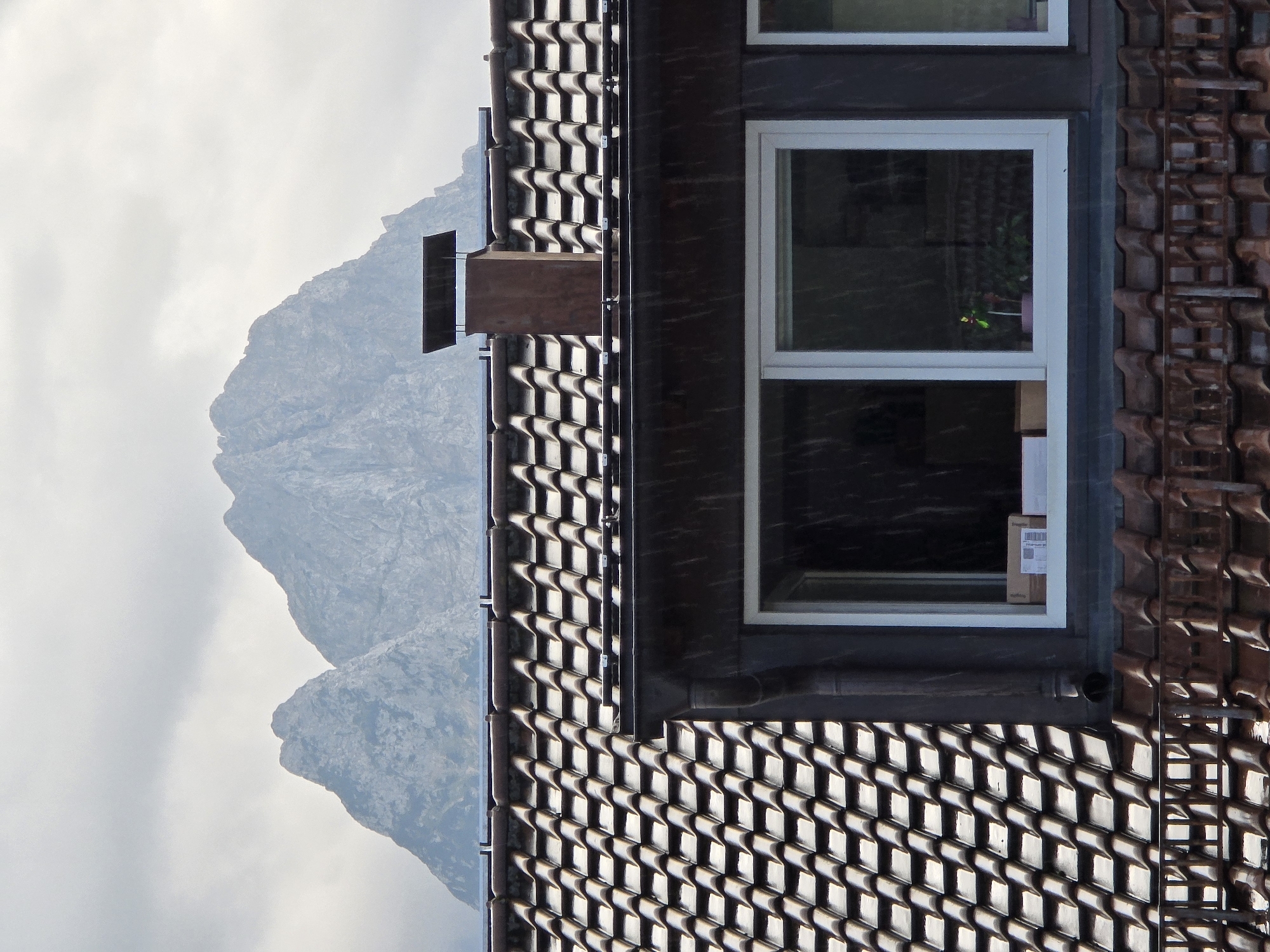 The image depicts a close-up view of a residential building's tiled roof and upper story window, with a mountainous landscape in the background. The roof is covered in dark, overlapping tiles, and a brick chimney with a metal cap stands prominently on the ridge. A large window with a white frame is visible on the upper floor, revealing the interior with boxes and a small potted plant with a yellow and green flower. Raindrops are visible falling throughout the scene, indicating inclement weather. In the distance, large, rugged mountains dominate the sky, their peaks partially obscured by clouds. The overall impression is one of a cloudy, rainy day in a mountainous region. While the image does not explicitly state its location, the mention of Garmisch-Partenkirchen, Germany, suggests this is a view of a building in or near that Bavarian town, known for its alpine scenery.