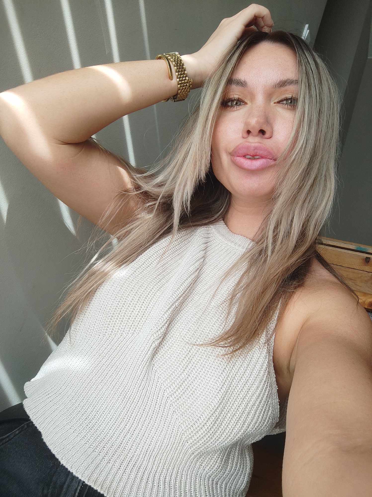 A fair-skinned woman is posing for a selfie indoors. She is seated with her left arm raised, her hand touching her hair, and a gold bracelet is visible on her wrist. She is wearing a white, knitted top and dark jeans or trousers. Light from a window is casting stripes across the wall behind her and also illuminates her arm. The background is partially visible, showing a gray wall and a glimpse of a wooden structure. The location is unknown as this photo doesn't have enough information to identify it as being taken in Votkinsk, Russia.