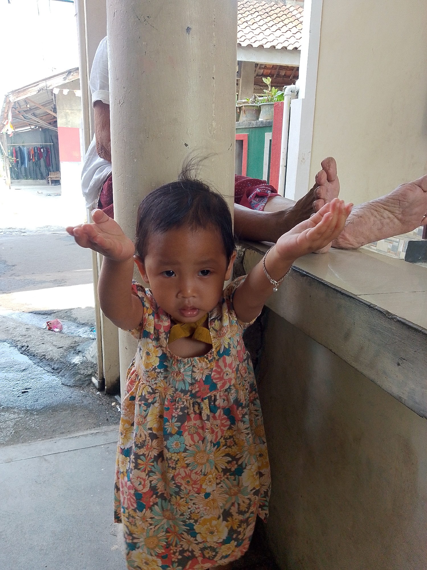 A young child in a floral dress stands in front of a concrete pillar, extending their arms forward with open palms. To the right of the child, two adult feet are visible, resting on a concrete ledge. The background reveals a building with a tiled roof and what appears to be an outdoor clothing drying area. The ground in front of the child is wet and shows signs of wear. The setting is likely a residential area in Bandar Lampung, Indonesia, possibly a porch or walkway.