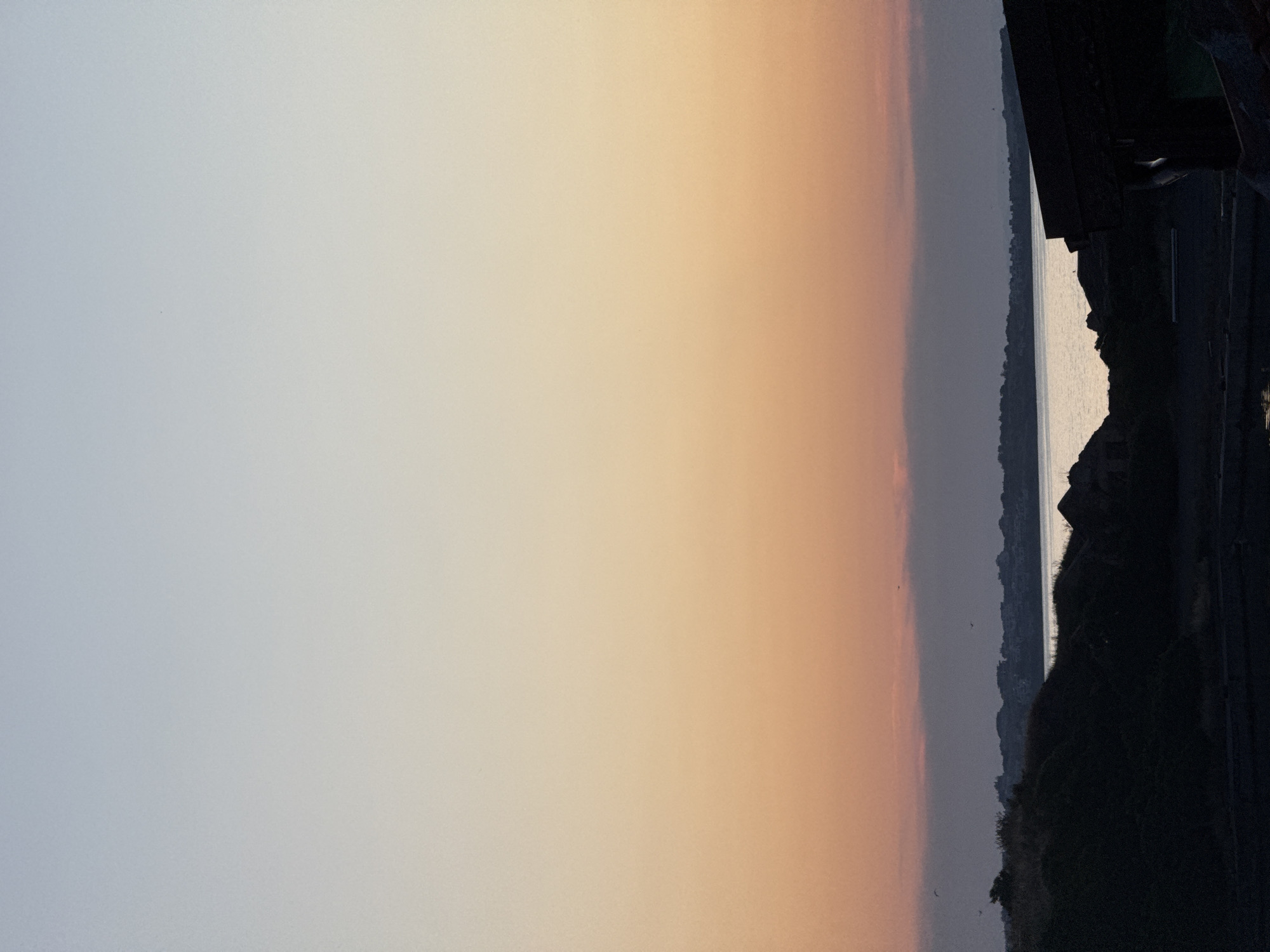 This image depicts a serene sunset scene over the Black Sea in Sozopol, Bulgaria. The sky transitions from a pale, cool blue on the left to a warm, peachy orange on the right, indicating the time of day is late afternoon or early evening. The sea is visible on the right side of the image, reflecting the soft hues of the sky.
A dark, silhouetted shoreline stretches across the mid-ground, suggesting a coastal landscape with some elevation, possibly cliffs. The land is covered in vegetation, creating a textured silhouette against the brighter sky and sea. In the foreground on the far right, there is a dark, structural element, possibly a building or a man-made overlook, also in silhouette. The overall mood is peaceful and tranquil, with the muted colors and quiet scene evoking a sense of calm. There are no visible people or text in the image. The weather appears clear, with no clouds obstructing the sunset. The location is identifiable as Sozopol, Bulgaria, known for its beautiful coastline and sunset views.