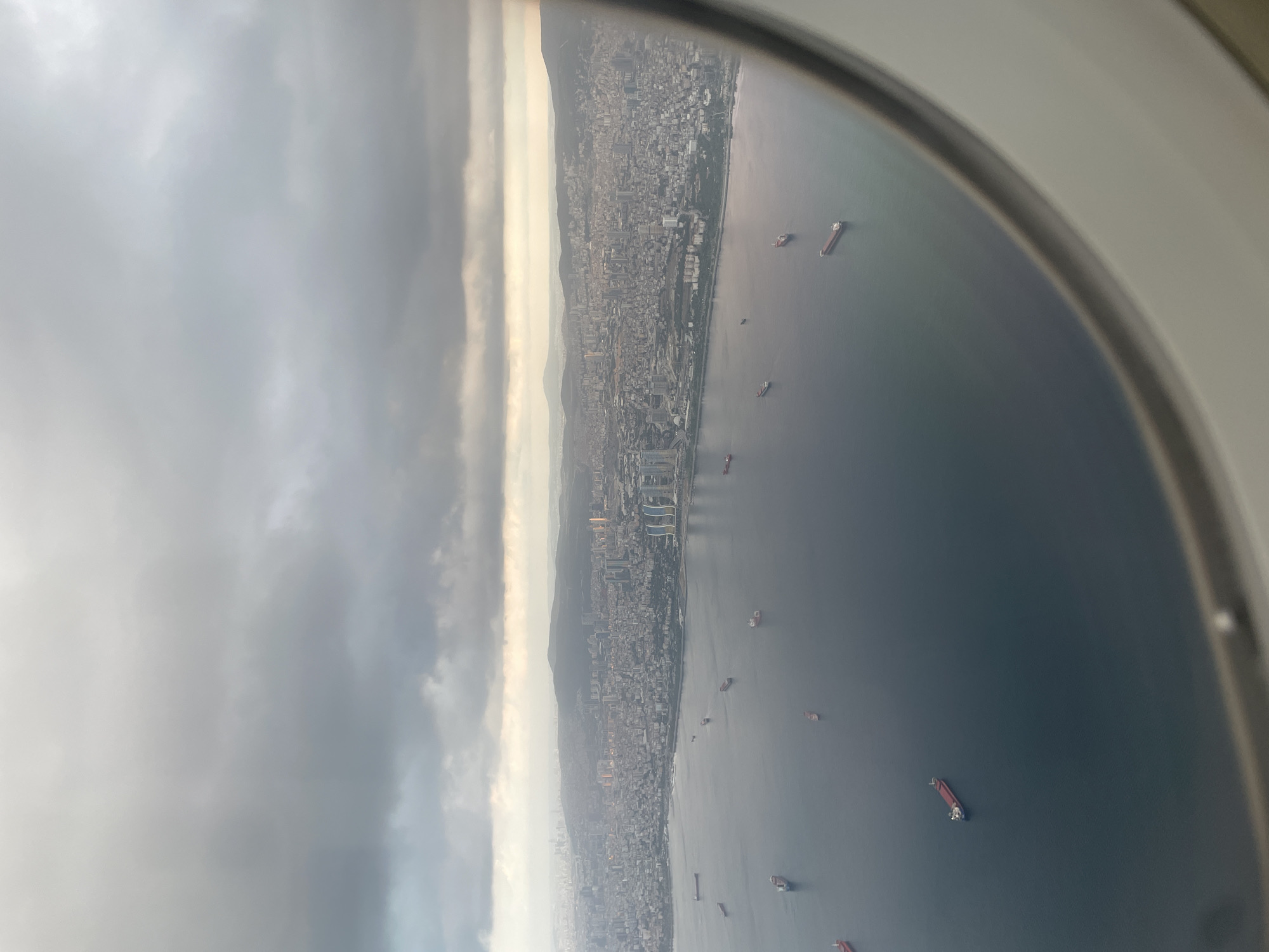 An aerial view captured from an airplane window shows a vast body of water in the foreground, dotted with numerous cargo ships of varying sizes. The water is a deep, calm blue. In the middle ground, a densely populated city stretches along the coastline. Buildings of different heights and styles are visible, creating a sprawling urban landscape. The city appears to be located on a bay or inlet, with the water meeting the land.
In the background, rolling hills or mountains are visible, fading into a hazy horizon. The sky is overcast with heavy, grey clouds, suggesting either an early morning, late evening, or a cloudy day. The lighting is muted and diffused, characteristic of such weather conditions. The overall impression is of a large coastal city with significant maritime activity under a cloudy sky. While the specific location is not identified, the architecture and the presence of the Baha'i House of Worship, or Flame Towers, strongly suggest Baku, Azerbaijan. However, the prompt states the location is Unknown, Türkiye. There are no people or specific actions clearly discernible in the image.