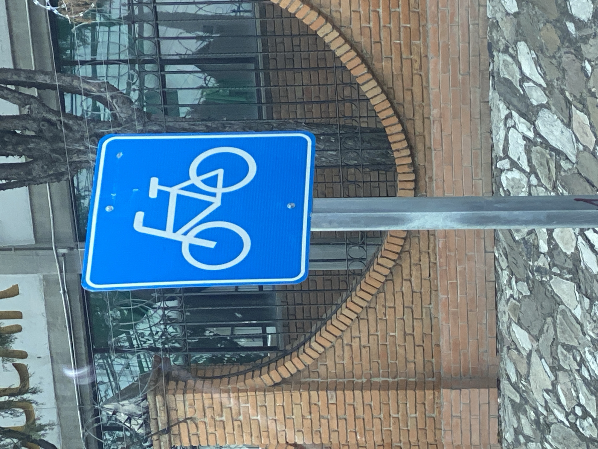 The image, rotated 90 degrees, features a prominent blue square sign with a white bicycle pictogram, indicating a cycling path or facility. The sign is attached to a metallic pole, extending from the right side of the frame.
Behind the sign, the background reveals an urban building facade. A significant portion consists of a red brick wall, characterized by a distinct curved arch also constructed from bricks. Further behind this arch and to the left, there is a reflective glass surface, likely a window or storefront, partially covered by a dark metal grille. Reflections of tree branches are visible on this glass.
To the right of the brick section, the building wall transitions to a different material: irregular, multi-toned grey and white stones, creating a textured, mosaic-like cladding. The overall lighting suggests it is daytime. No people or specific activities are depicted, portraying a static scene of urban infrastructure and architecture. The bicycle sign itself contains no text, relying solely on the universally recognized pictogram. A faint, indistinct golden letter or symbol is barely visible on another cropped sign on the far left.