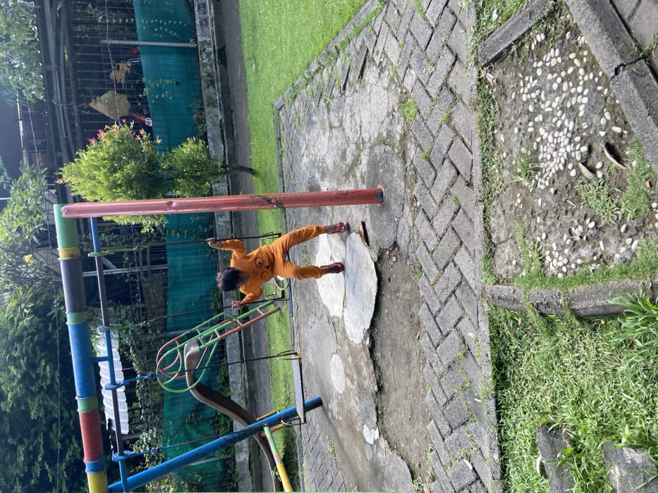 A young girl in an orange shirt is swinging on a swing set in a small park. The swing set is made of metal and has a slide attached to it. The girl is wearing red shoes and has her hair pulled back. The park is located in Kecamatan Sidoarjo, Indonesia. The ground is made of brick and the area around the swing set is covered in grass. There are plants growing in the park. The girl is swinging back and forth. The swing set is located near a fence. There is a large green area behind the fence. The area appears to be a residential area.