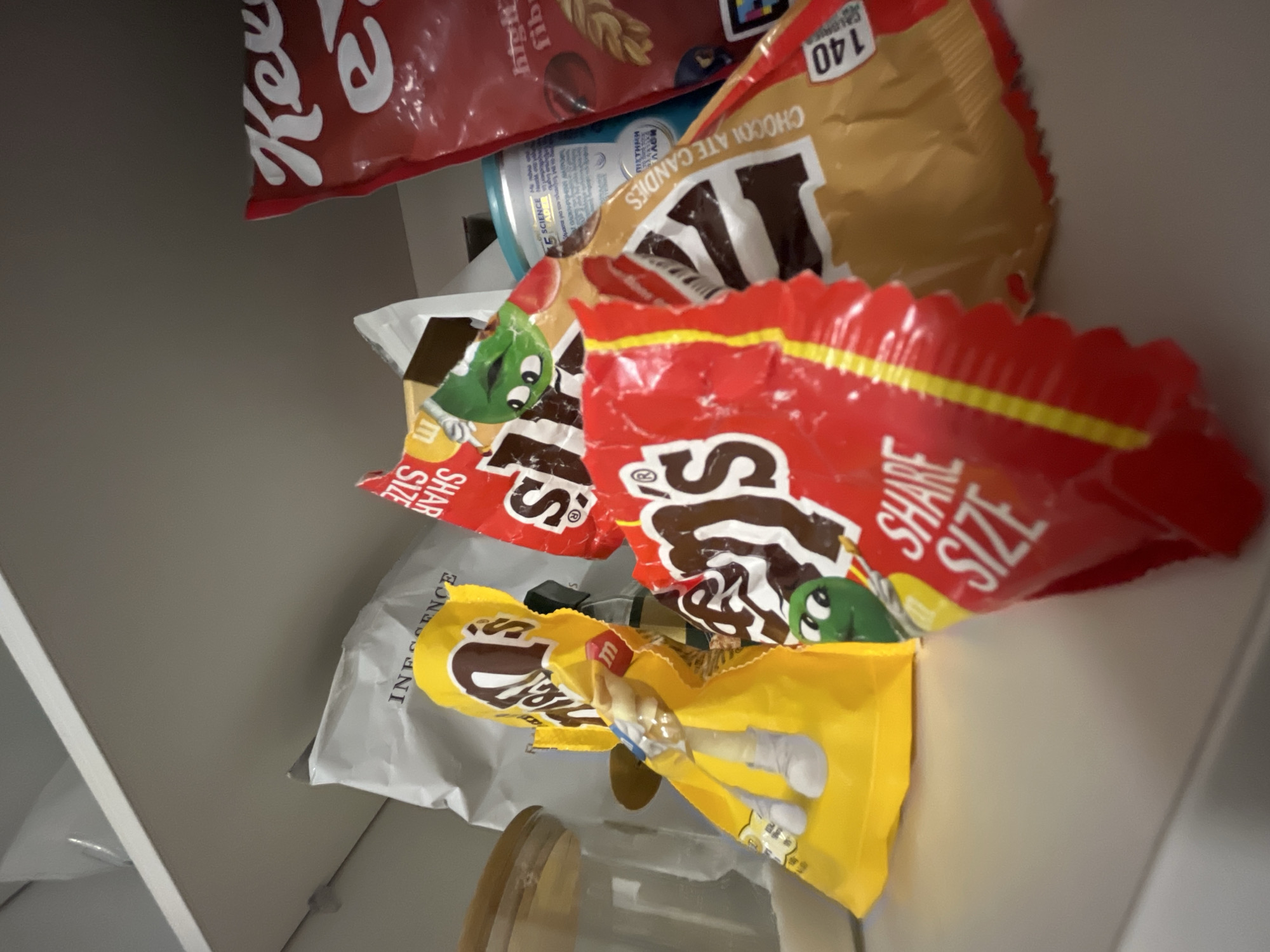 This image depicts a close-up, low-angle shot of several bags of M&M's candy, along with another red bag, possibly of a cereal, on a white shelf. The M&M's bags are in various states of being open and crumpled, with some featuring the iconic M&M's characters. One prominent bag is bright yellow with a white "M&M's" logo and a character illustration. Another is red and white, also with the M&M's branding and the words "SHARE SIZE". A brown bag with M&M's branding is also visible, partially obscured by the red bag. Behind these, a large red bag with white lettering, possibly "Kellogg's", and some text in a foreign language, is visible. A portion of a blue cylindrical container with text and graphics is also present. The shelf appears to be indoors, likely in a pantry or on a cabinet. There are no people visible in the image. The image does not contain any information or context that would place it in Limassol, Cyprus.