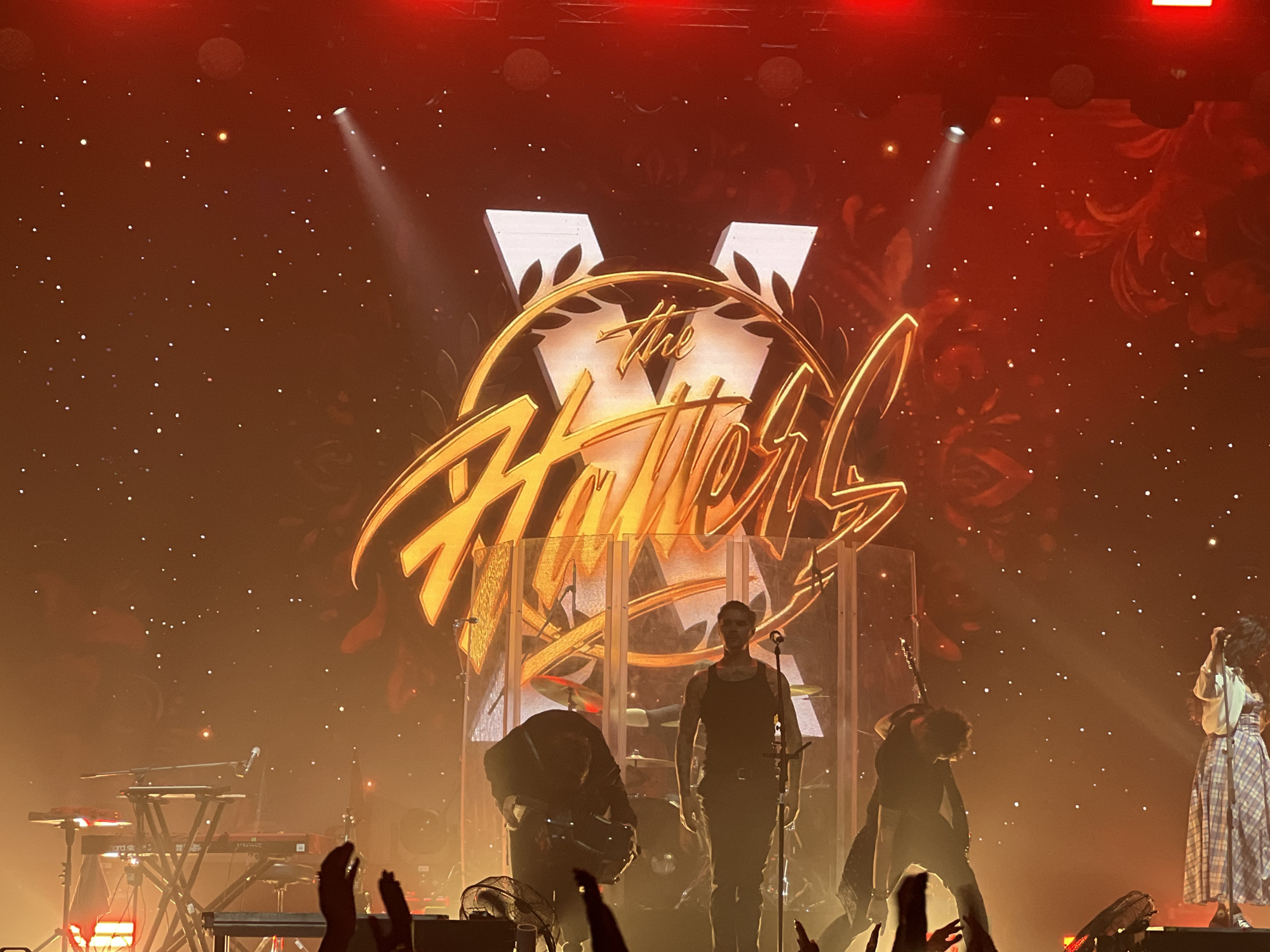 The image shows a concert stage, likely in Izhevsk, Russia, given the band "The Hatters" performing. The band's logo is displayed prominently in the background. Several band members are on stage, including one wearing a black tank top standing center stage. A woman in a long patterned dress and white top stands to the right. Other band members are further back, either performing or adjusting instruments. A keyboard setup is visible on the left. The stage lighting is orange and red, creating a warm atmosphere. Many hands of audience members are reaching up in the foreground.