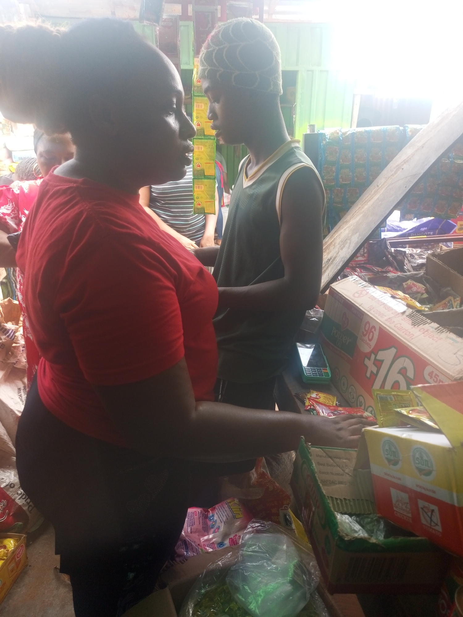 In a brightly lit indoor market in Sabon Tasha, Nigeria, a woman in a red shirt and black pants stands at a stall, interacting with a young man wearing a green vest and patterned head covering. The stall is filled with packaged goods, including a large box labeled "Power Oil +16 96 Sachets" and smaller packages marked "SOL". A green and black phone rests on the stall's surface amidst the goods. Other customers and market vendors are visible in the background.