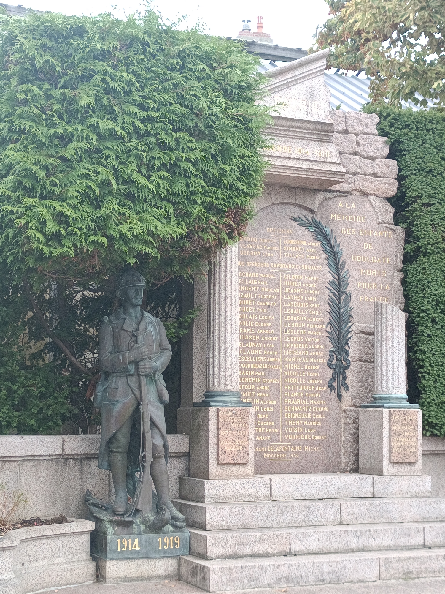 This is a photograph of a war memorial in Houlgate, France. The memorial is made of granite and features a statue of a soldier standing at attention. The soldier is wearing a helmet and a uniform and is holding a rifle. The base of the statue has the dates "1914" and "1919" inscribed in gold. Behind the statue is a large granite slab with names inscribed on it. The names are listed in columns and are accompanied by a laurel wreath design. To the right of the slab, there is a pillar with more inscriptions. The memorial is surrounded by lush green bushes and trees. The sky is overcast.