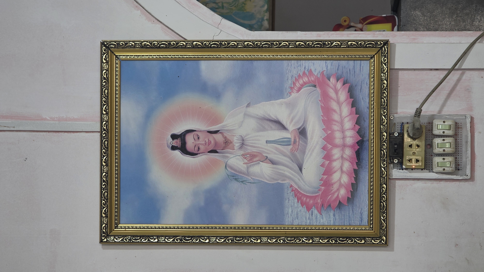 A framed print hangs on a pale pink and white wall. The frame is gold with decorative patterns. The print features a woman in white robes with a lotus flower. To the right of the print is an electrical outlet with a plug and a row of three switches. The scene is located in Nha Trang, Vietnam.