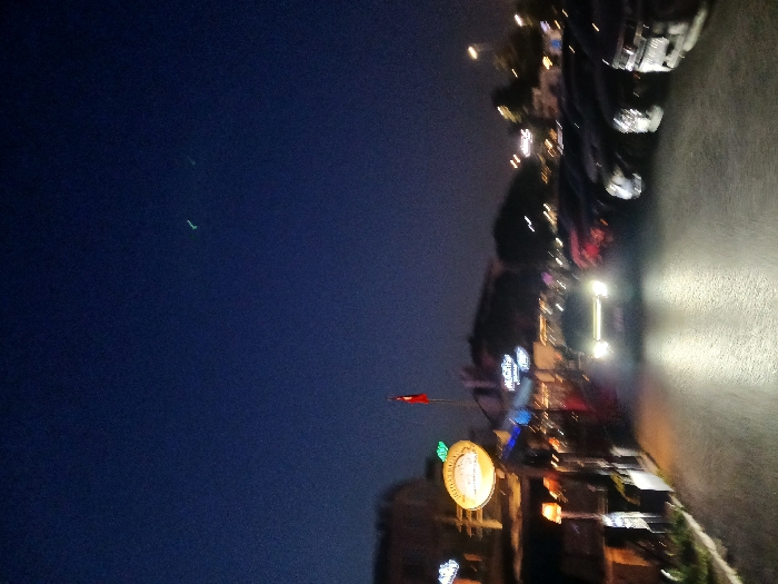 The image shows a street scene at night in Istanbul, Turkey, specifically at the coordinates 40.956 latitude and 28.821 longitude. The scene is dark, with a few streetlights illuminating the road. There are several cars parked on both sides of the road. A few individuals are walking on the sidewalk in the distance. The camera appears to be positioned inside a vehicle, facing forward toward the street. The street is paved and has streetlights on either side. There is a Turkish flag on a pole in the foreground, and a large orange sign that is illegible. The sky is dark blue with two small bright lights. The image is taken from a street in Istanbul, Turkey. The cars are parked and illuminated by streetlights. It is evening, with a clear dark sky, and the road is paved. The image is blurred, suggesting it was taken from a moving vehicle.