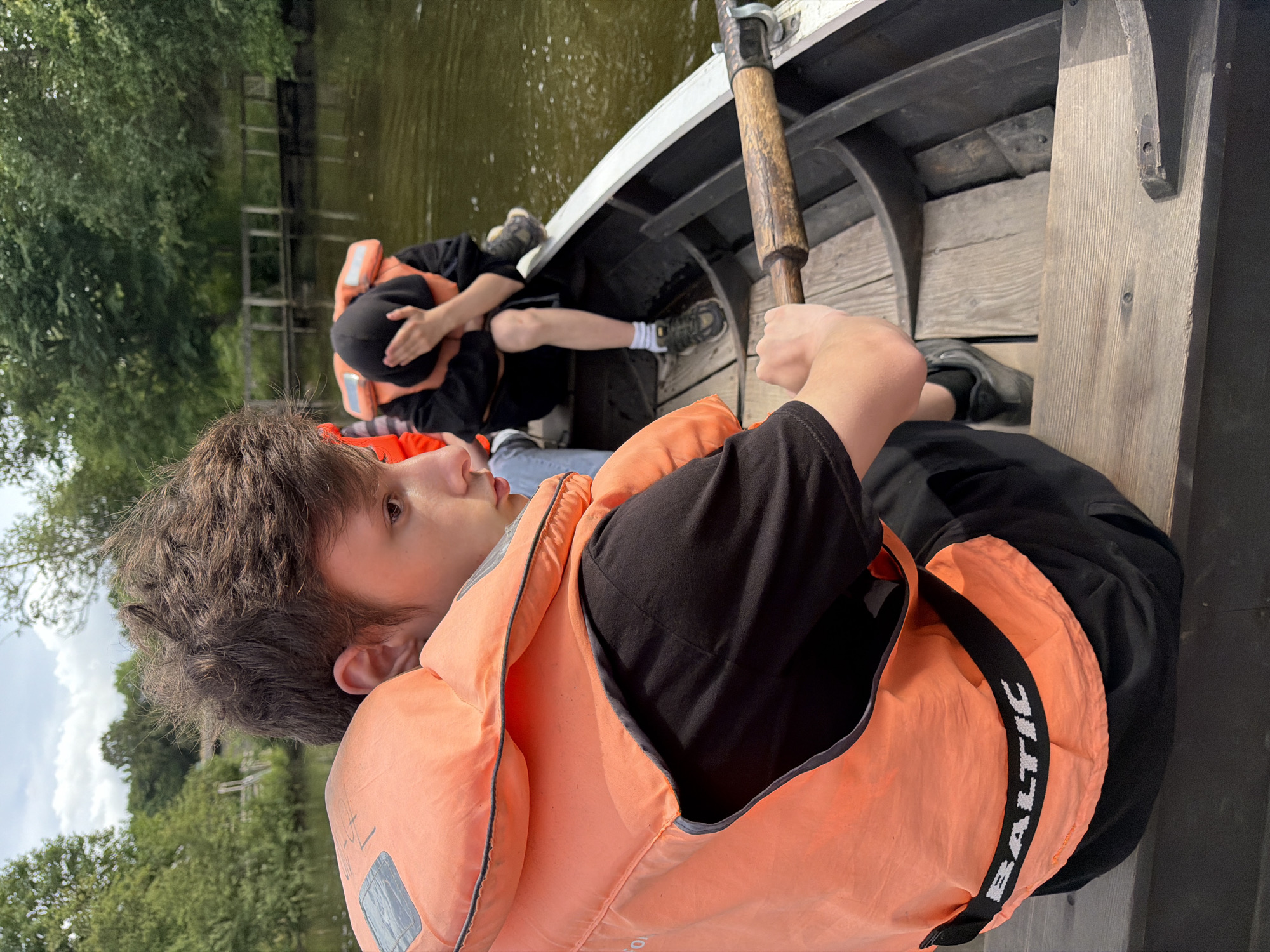 In the city of Allerslev, Denmark, two individuals are seated in a wooden rowboat on a body of water. The individual in the foreground, facing away from the camera, wears an orange life vest over a black t-shirt and black shorts, with the brand "BALTIC" visible on the vest. This person's hands are gripping an oar, positioned for rowing. The second individual, further back in the boat and partially obscured, also wears an orange life vest and a dark hooded garment, with their face covered by their hand. The environment features a tranquil body of water, with lush green trees and vegetation lining the banks. In the background, a wooden bridge or walkway is visible extending over the water. The sky above is partly cloudy.